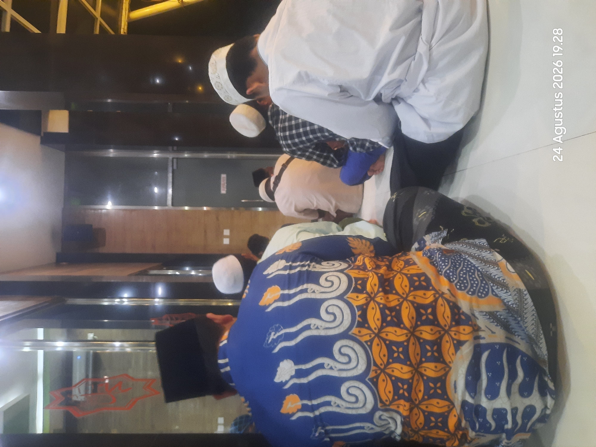 The image shows a group of men sitting on the floor, facing away from the camera, likely in prayer. They are wearing traditional Islamic attire, including peci caps and long shirts. One man in the foreground wears a vibrant blue batik shirt with an intricate orange and white pattern. Another man to his right wears a white peci and a white robe. The men are seated on a light-colored floor, possibly in a mosque or prayer hall. In the background, a reflective surface, likely glass, displays a red logo. The environment appears to be indoors with modern architecture, suggested by dark pillars and lighting fixtures. A timestamp in the lower right corner indicates "24 Augustus 2026 19.28." The specific location in Sukokerto, Indonesia, cannot be determined from the visual information alone, as there are no identifiable landmarks or signs.