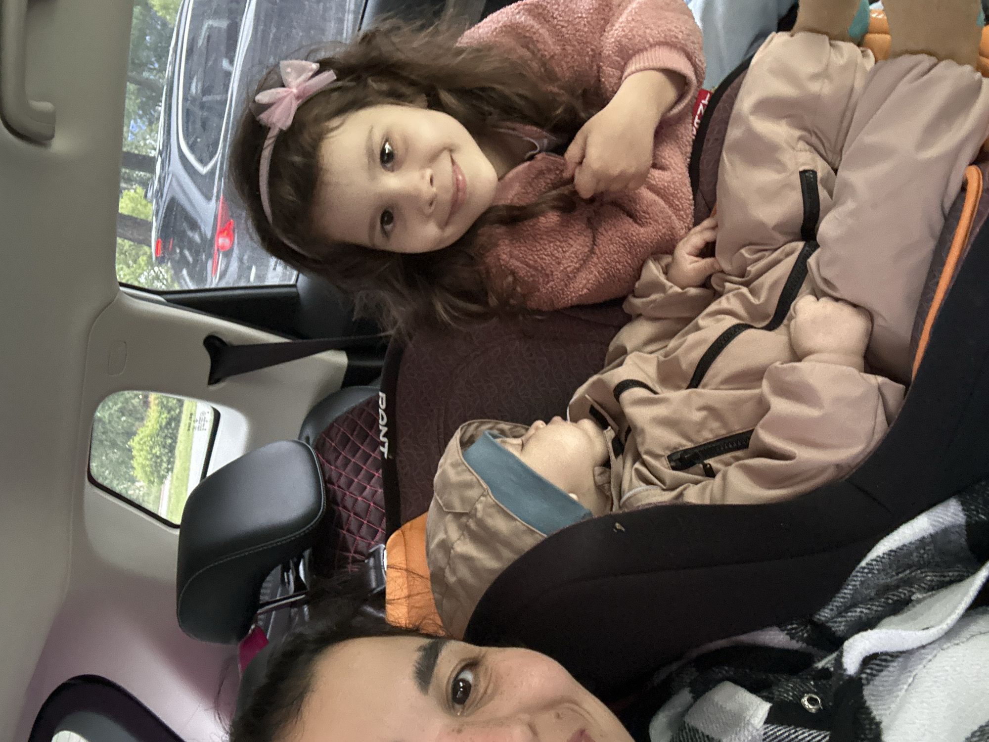 This image, rotated 90 degrees clockwise, captures a moment inside a car, likely a selfie. In the upper part of the frame, a young girl with long brown hair, adorned with a pink bow headband, smiles brightly at the camera. She is wearing a textured pink sweater. Below and to her right, another child, clad in a light beige hooded jacket with a blue lining, appears to be resting or asleep, positioned on what looks like a car seat or booster.
The bottom right corner of the frame shows the partial face of an adult, looking directly at the camera, wearing a black and white plaid shirt. The car's interior is visible, including a dark headrest and a red and black patterned car seat or cover on which the text "PANT" is visible. Through the car window in the background, a dark vehicle is partially seen, along with green trees and foliage, suggesting a daytime setting with natural light. The overall mood is cheerful, primarily due to the young girl's happy expression.