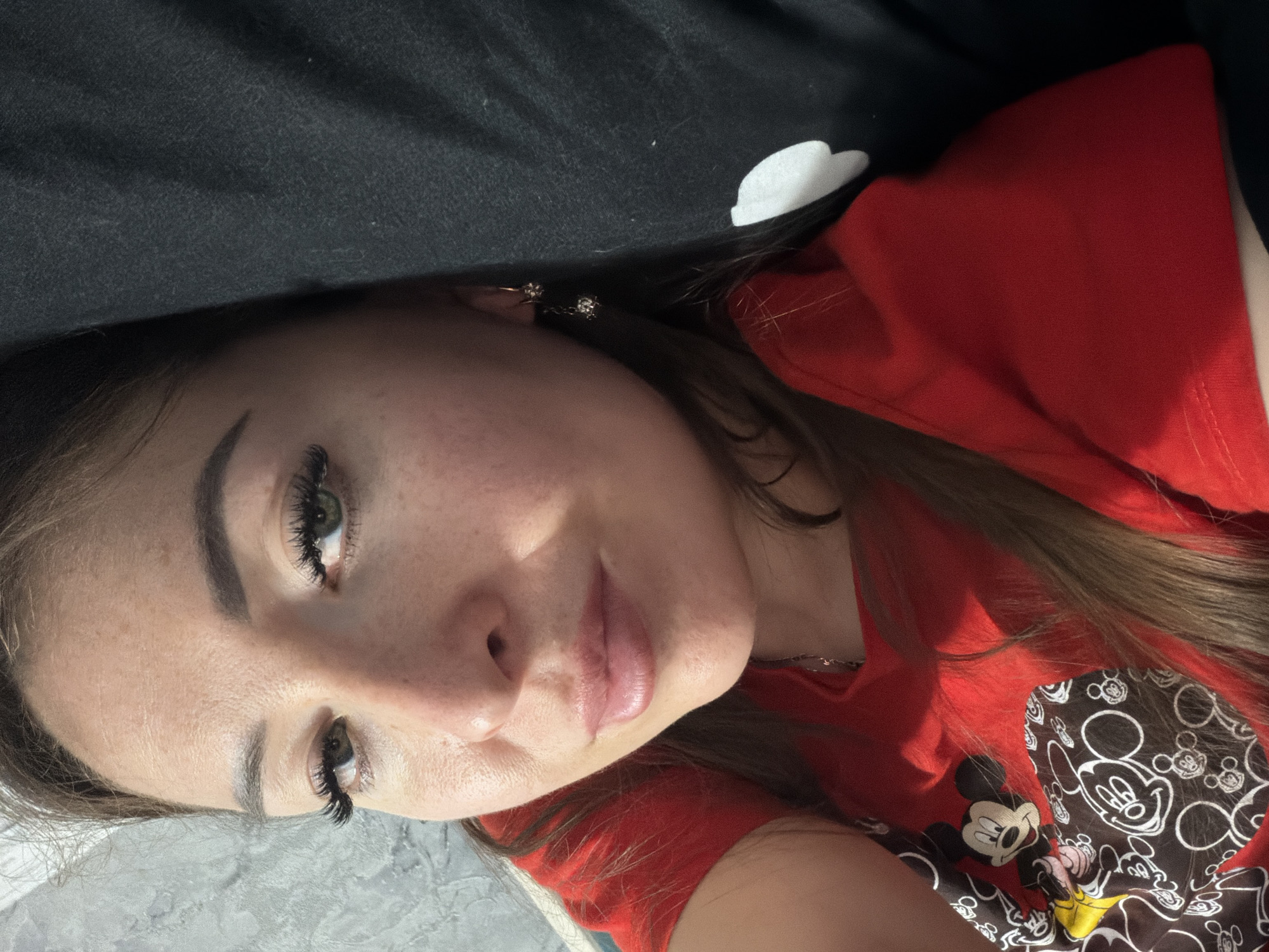 This media file is a close-up, low-angle, portrait-style selfie of a young woman lying down. She is wearing a red t-shirt with a Mickey Mouse print. The background consists of a black fabric surface with white polka dots and a textured gray surface. Her expression is relaxed and she appears to be in a comfortable, indoor setting. The lighting suggests it is daytime. No visible text is present in the image. The location context is Япрыково, Russia.