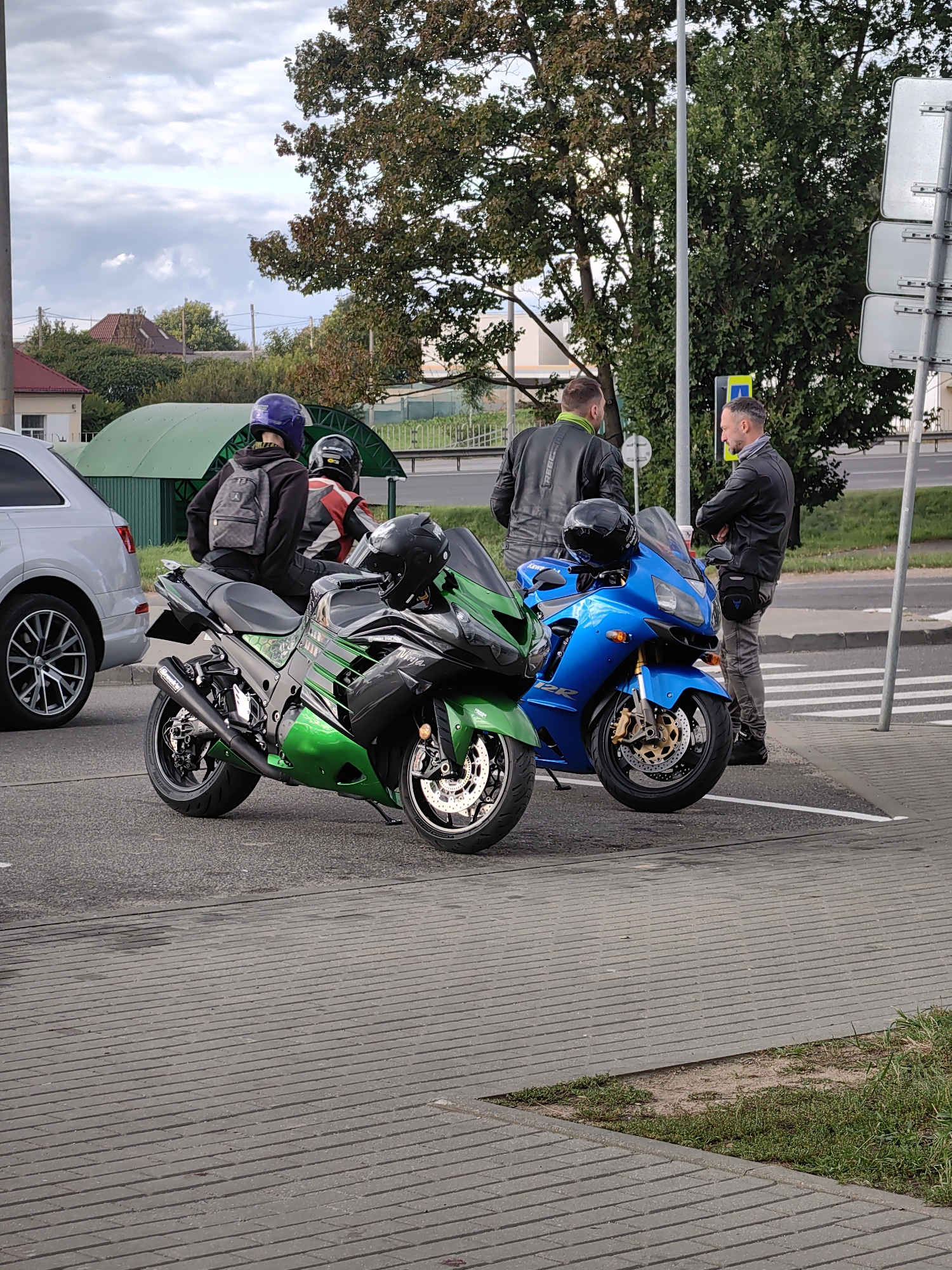 Two motorcyclists are standing near their motorcycles on a paved area adjacent to a street. The motorcycle on the left is green and black, a Kawasaki Ninja, with a person wearing a blue helmet and a backpack sitting on it. The motorcycle on the right is blue, with a person standing next to it. Both individuals appear to be adult males. One man is wearing a black leather jacket and grey pants, with a fanny pack around his waist. The other man is wearing a black leather jacket and also appears to be wearing grey pants. They are standing on a sidewalk or paved area with a pedestrian crossing visible in the background. Trees and buildings are visible in the background, suggesting an urban or suburban environment. The sky is cloudy. The location is identified as Фаніпальскі сельскі Савет, Belarus.