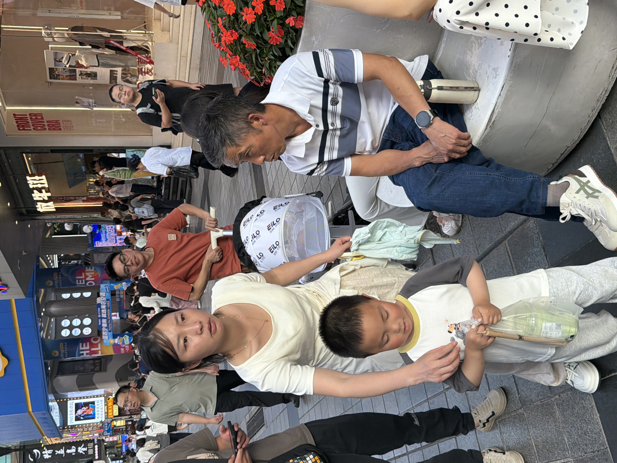 This is a street-level, medium shot photograph taken in Yuzhong District, China, likely in an urban commercial area with pedestrian traffic and storefronts. In the foreground, a young boy wearing a gray and white t-shirt and light gray sweatpants is holding a clear plastic bag containing a drink and other items. His mother, a woman with dark hair and a light-colored dress, is reaching towards the boy, her hand visible near his. Behind them, a man with short gray hair is seated on a white bench, wearing a striped polo shirt and blue jeans, and looking to his left. A white thermos is on the bench next to him. To the left of the seated man, another man in a white and gray striped shirt is also looking to his left. In the background, a diverse crowd of people are walking along a busy street lined with illuminated signs and storefronts. A baby stroller is visible between the woman and the seated man. The overall scene depicts a casual moment in a bustling city environment.