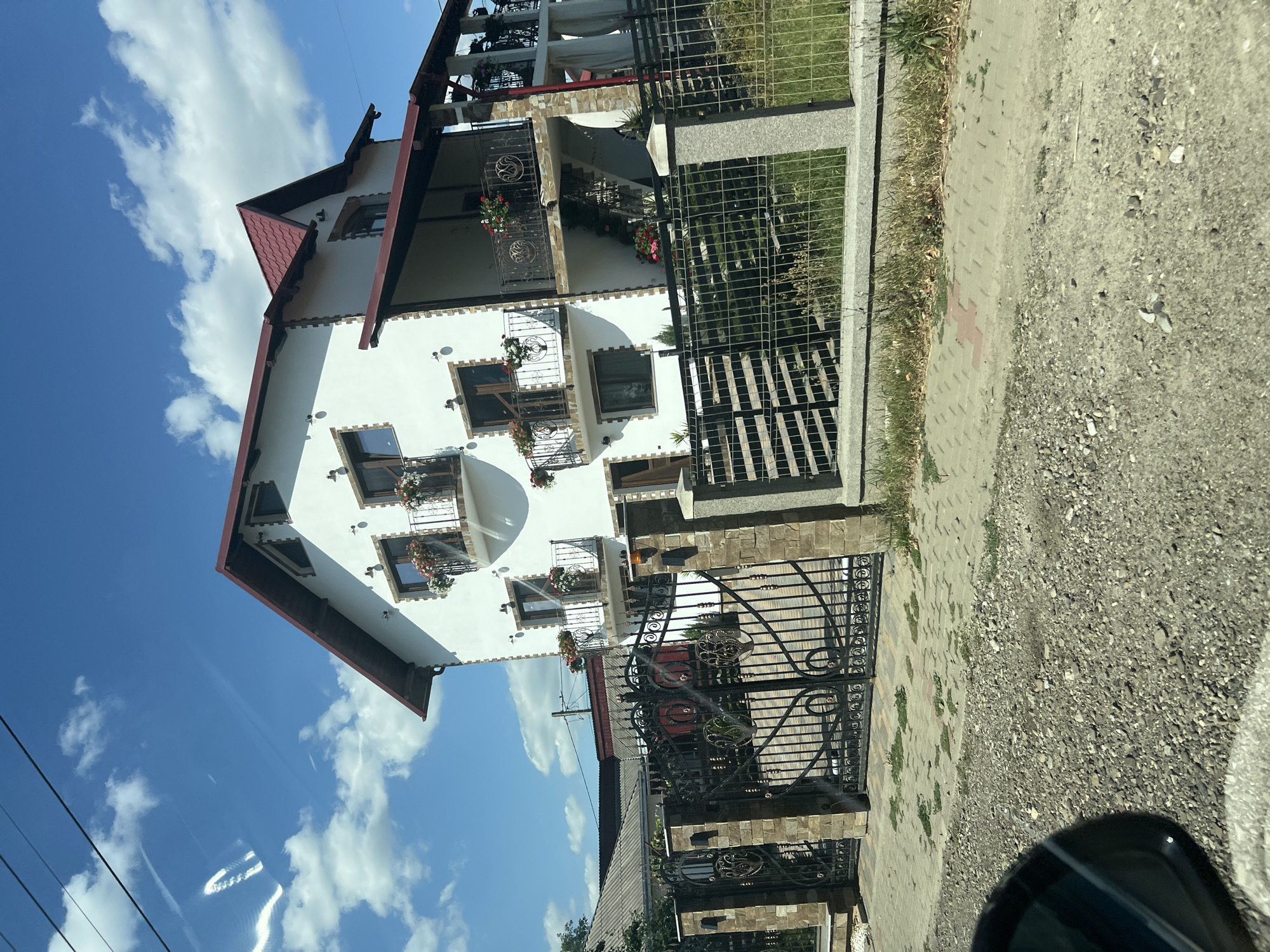 The image depicts a white, multi-story house with a red-tiled roof, located in Cornu Luncii, Romania. The house features multiple balconies with decorative railings and flower boxes. A large, ornate black metal gate, supported by stone pillars, marks the entrance to the property. A metal fence runs along the perimeter, with a stone border and grass. The scene is set against a bright blue sky with scattered white clouds. The foreground shows a gravel and paved pathway. There are no people visible in the image.