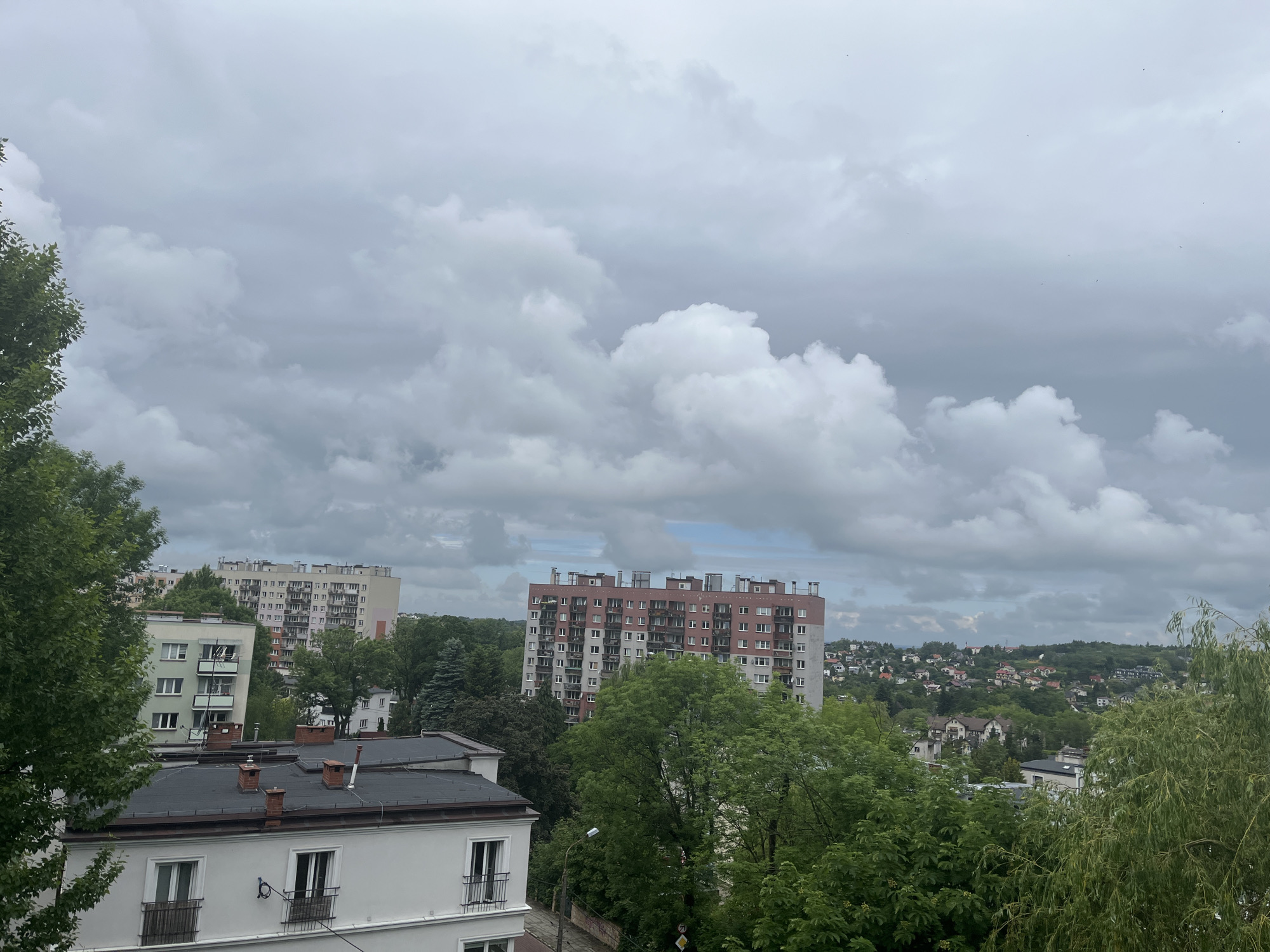 The image presents a wide, high-angle view of an urban or suburban landscape under an overcast sky. The foreground features the roof and upper facade of a white building with dark eaves, multiple chimneys, and windows with railings. A dense canopy of green trees frames the left side of the scene and extends throughout the middle ground.
In the midground, several large apartment blocks dominate the view. On the left, a light-colored, multi-story apartment building is partially visible, featuring numerous balconies. To its right, a prominent, tall apartment complex with a distinctive reddish-brown and gray facade stands out, also adorned with many balconies and visible rooftop structures. Lush green trees surround these buildings, with some obscuring parts of their lower sections.
Beyond the immediate urban structures, the landscape recedes into a series of green hills dotted with a cluster of smaller residential houses, indicating a sprawling community or suburb. The sky above is entirely covered by heavy, gray and white clouds, suggesting an overcast day with diffused light. A few tiny, dark specks, possibly birds, are faintly visible against the clouds in the upper right quadrant. No people or specific activities are discernible, and there is no visible text. The scene depicts a calm, cloudy day over a densely built environment integrated with significant natural greenery.