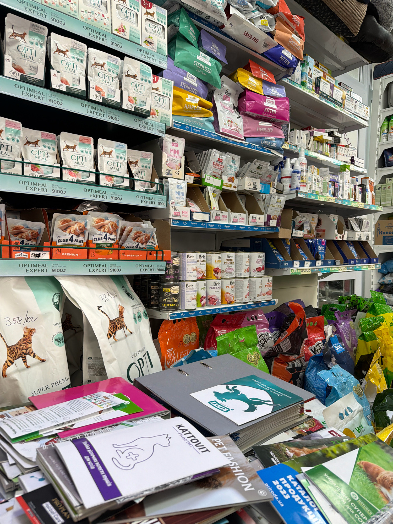 The image depicts a retail setting, likely a pet supply store, with shelves stocked with various pet food products. The merchandise includes bags of dry pet food, pouches of wet food, and canned food, with brands like "OPTIMAL EXPERT," "CLUB 4 PAWS," and "Brit Care" visible. The lighting suggests an indoor environment, and the overall presentation of the products indicates a commercial establishment. There are no people visible in the image. The presence of Cyrillic text on some of the packaging, such as "СЕРВІС ВЕТ-РЕГІОН" and "КАТАЛОГ 2023," strongly suggests the location is in Ukraine, and likely Kyiv, as it is the capital and largest city.