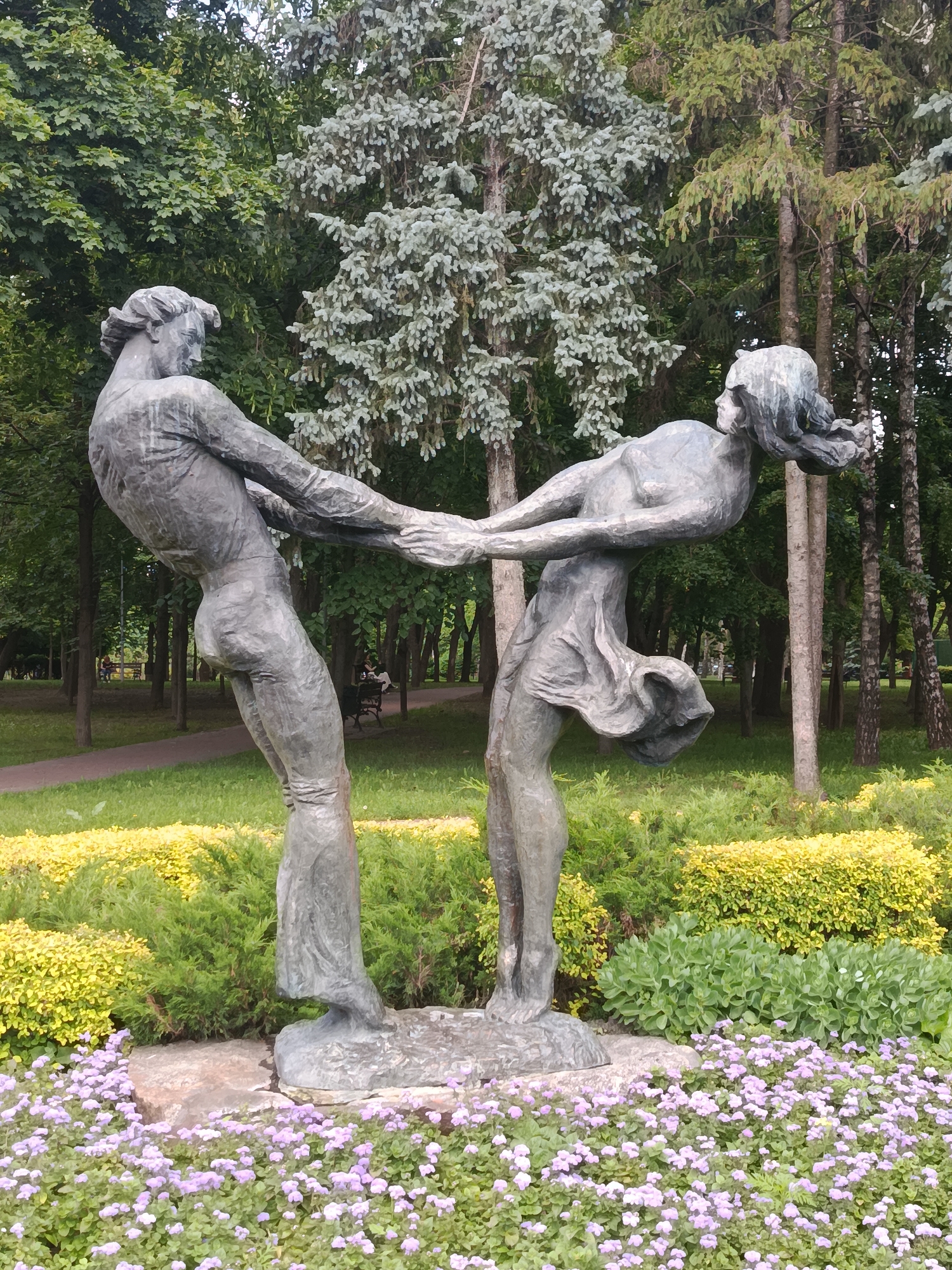 A full shot captures a bronze sculpture of a man and woman in a park. The man is on the left, facing the woman and holding her hands. He is leaning back, with his left leg bent and his right leg extended. The woman is on the right, facing the man and holding his hands. She is leaning back, with her left leg bent and her right leg extended. Her dress is flowing as if in motion. They are both in a dynamic pose, appearing to be dancing or in mid-movement.
The sculpture is positioned in front of a lush green garden with a variety of plants. In the foreground, there is a bed of small purple flowers, which are in full bloom. Behind the sculpture, there are manicured hedges with yellow and green foliage. Further in the background, a path winds through a grassy area, bordered by trees. The trees are tall and varied in species, with some having light green, needle-like leaves and others having broader, darker green leaves. In the distant background, out-of-focus figures can be seen sitting on benches, suggesting a public park setting. The sky is not visible in the frame. The overall impression is one of a serene and artistic outdoor space.