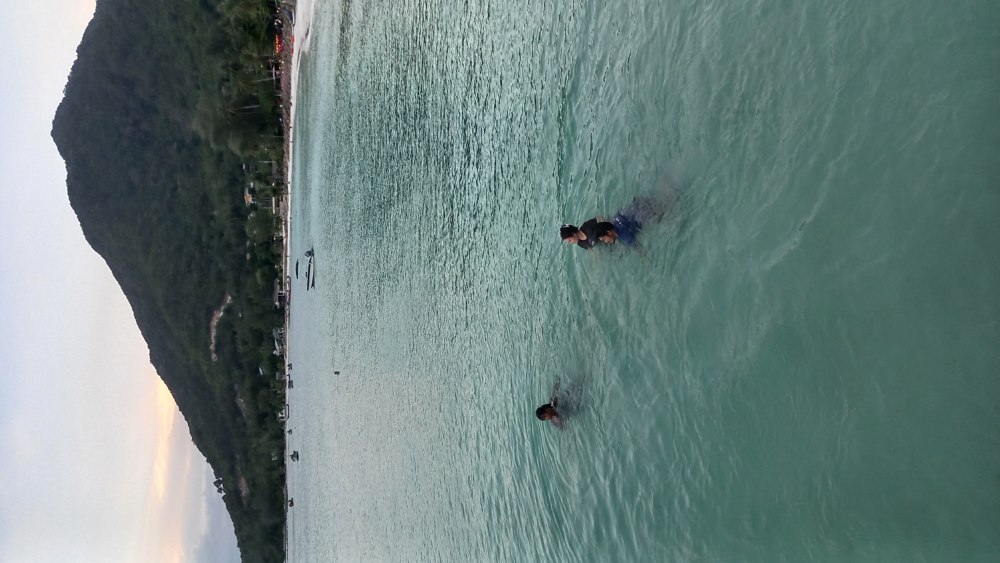 In the waters of Ko Pha-ngan District, Thailand, at least four individuals are visible swimming. In the foreground, a group of three people are together, with one person appearing to be submerged up to their neck and another person swimming near them. Further out in the water, a fourth individual is swimming. The sea is a clear, greenish-blue, with gentle ripples on the surface. In the background, a white sandy beach stretches along the coastline, lined with palm trees and buildings. A large, green, forested mountain rises behind the beach, its slopes covered in dense vegetation. Several boats are anchored offshore, and in the distance, the sky is a pale gray, with hints of sunset colors near the horizon. The overall scene depicts a serene coastal environment with recreational activity.