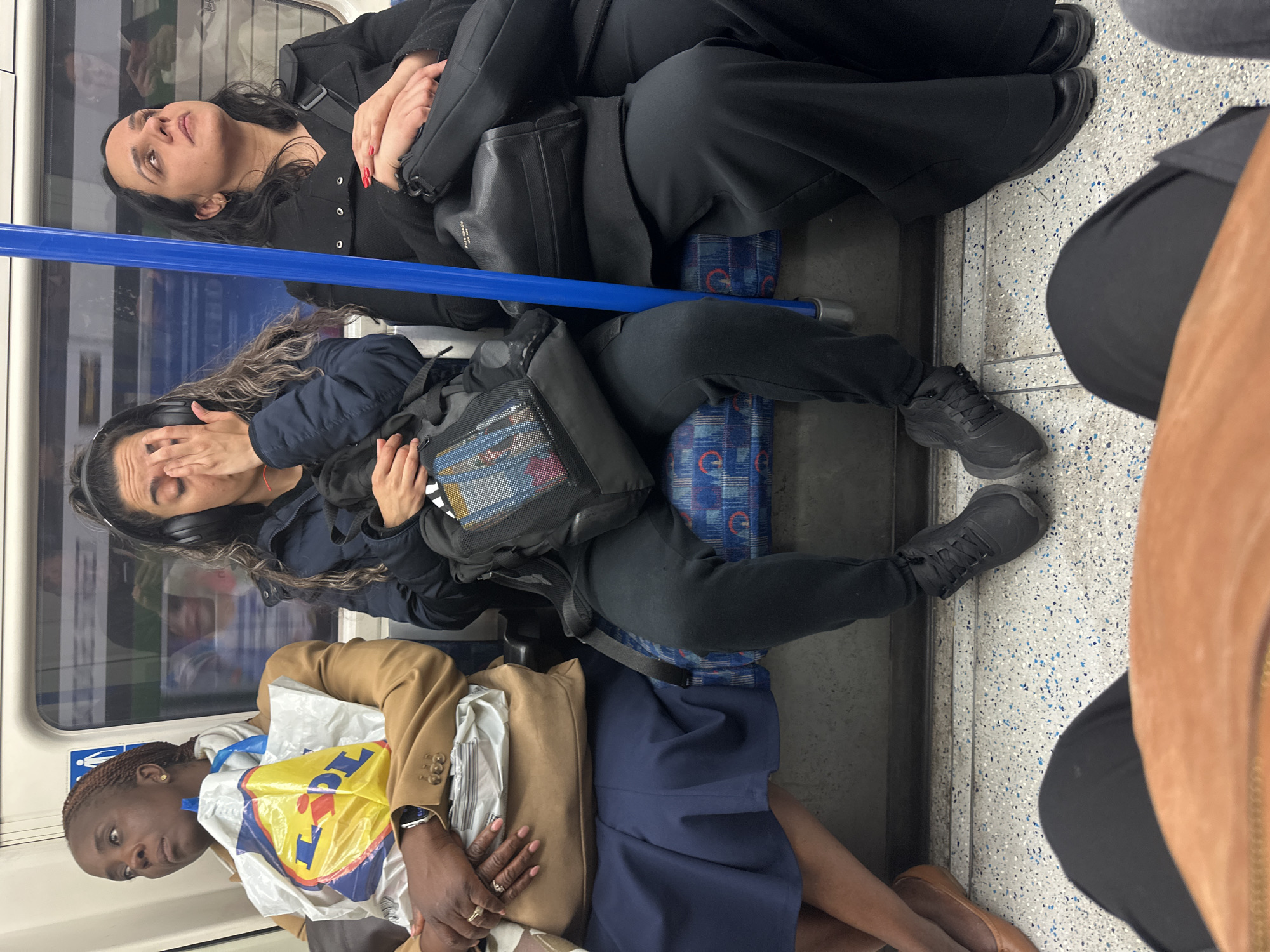 Three women are sitting on seats in a London Underground train carriage.
The woman closest to the camera is Black, with braided hair and is wearing a beige blazer, blue skirt, and carrying a yellow and white Lidl shopping bag. She has a wristwatch on her left wrist.
The woman in the middle is wearing headphones, a dark blue jacket, and black pants. She is holding her hand to her face. She is carrying a black backpack.
The woman farthest from the camera is wearing a black coat and black pants. She has red nail polish. She is carrying a black handbag.
The train carriage has blue patterned seats and a blue handrail. A window is visible behind the women. The train interior is clean. The image has been rotated.