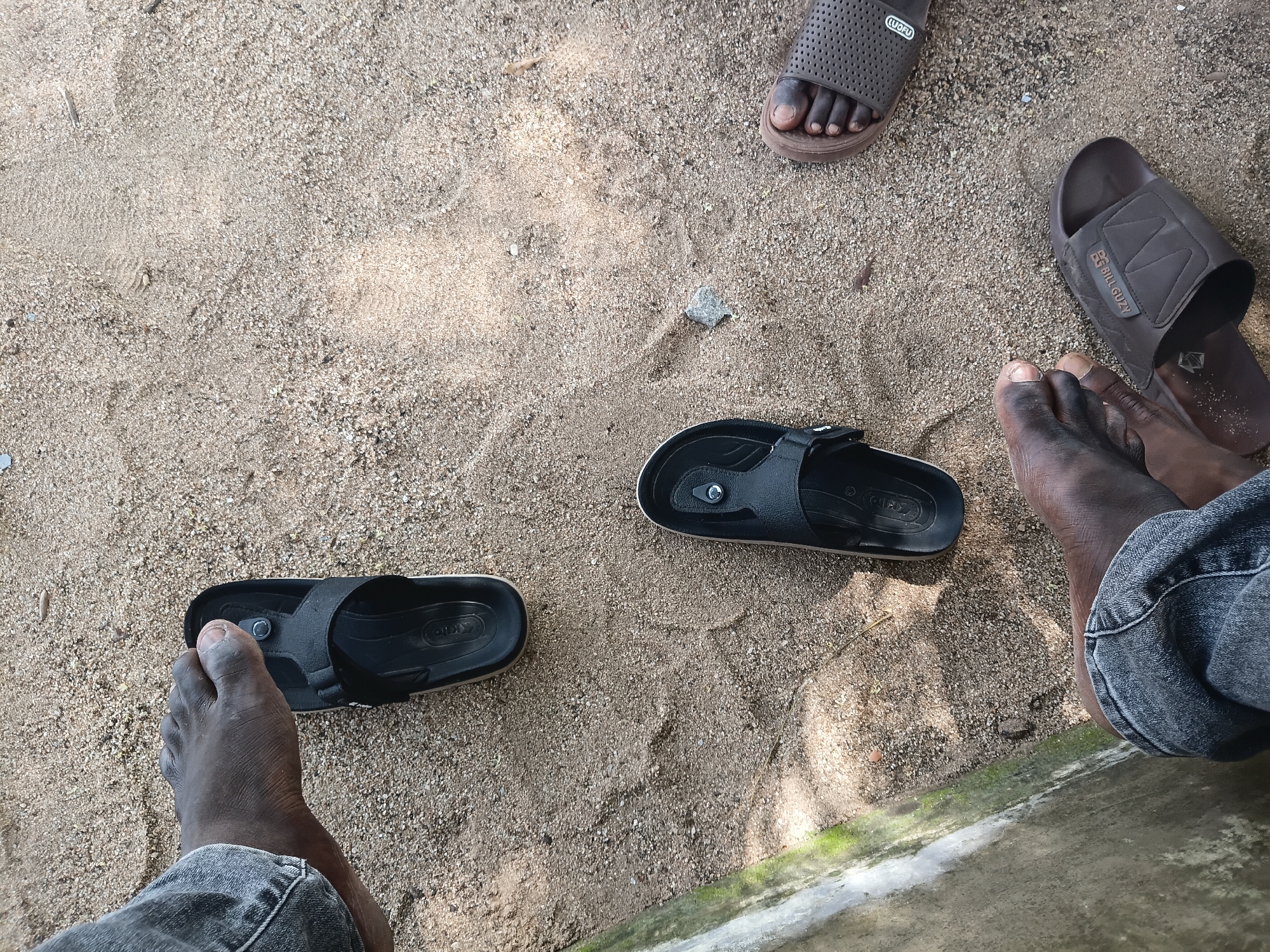 The image shows a close-up overhead view of several pairs of feet and sandals on a sandy surface, likely outdoors. The sandy ground is coarse, with small pebbles and some debris. In the upper left corner, a left foot, clad in a grey denim jean, is visible, with a black sandal placed over the toes. Below and to the right of this, another black sandal lies open. Further down, a brown sandal with a perforated design is seen on a right foot. In the lower right, another brown sandal, with the branding "BILL GUZY" visible, is on a left foot. A concrete structure, partially covered in moss, borders the left side of the image, suggesting an outdoor area possibly near a building or pathway. The lighting suggests it is daytime, with some shadows cast on the sand. The setting is consistent with an outdoor location in Jalingo, Nigeria, such as a backyard, park, or roadside, where people might be casually dressed.