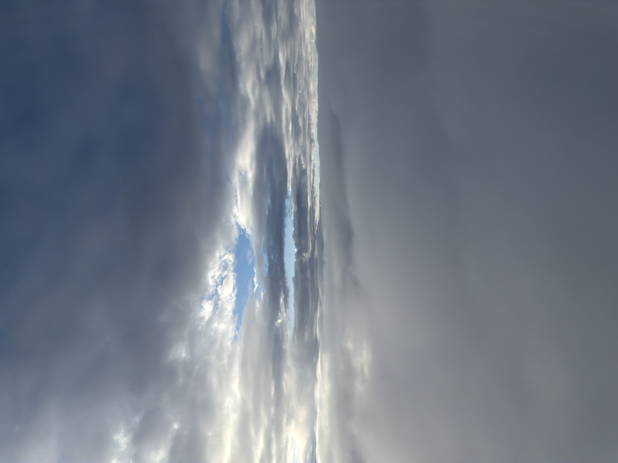 The image depicts a view from an aircraft, looking upwards at a cloudy sky. The clouds are layered, with darker, heavier clouds dominating the upper portion of the frame and lighter, wispy clouds towards the horizon. A patch of bright blue sky is visible through a gap in the clouds in the upper middle section of the image. Below these clouds, a dense layer of white, fluffy clouds fills the lower half of the frame, obscuring any view of the ground. The lighting suggests it is daytime, likely during overcast conditions. The location is specified as Valle, Norway, though this is inferred from contextual information rather than visual cues within the image itself, as the scene is entirely celestial. There are no people, subjects, or discernible objects in the image apart from the clouds and sky.