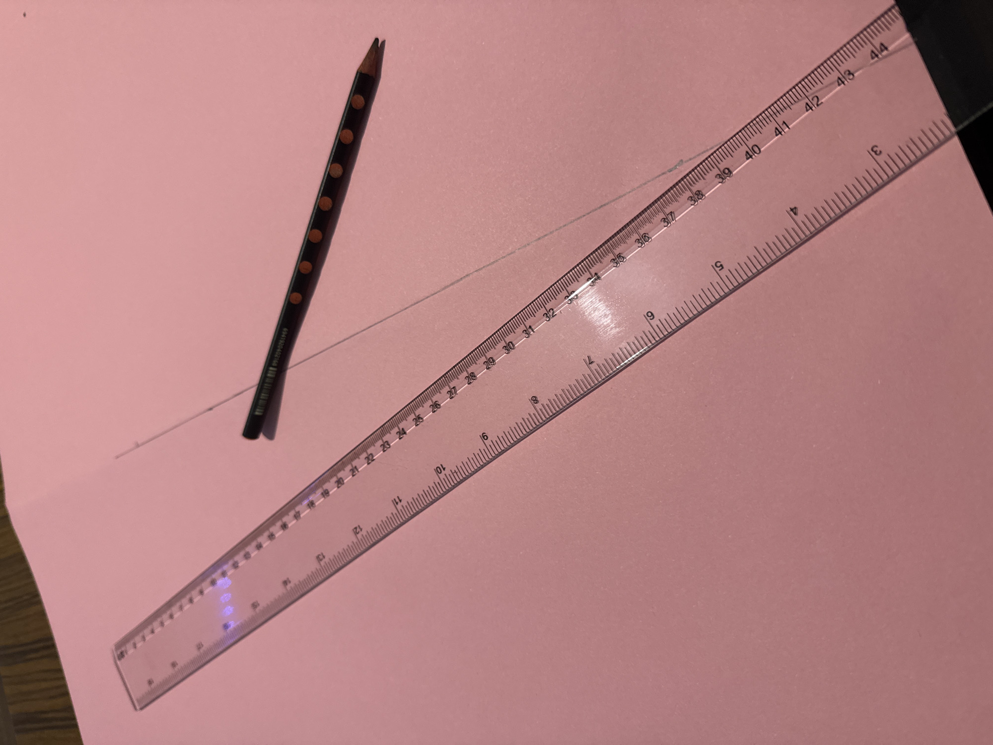 An overhead shot captures a dark pencil and a transparent ruler laid diagonally across a pink sheet of paper, suggesting an activity of drawing or measuring. A faint gray line is already visible on the paper, extending from under the pencil tip to near the ruler.
The pencil, positioned in the upper left, is dark brown or black with several evenly spaced orange-brown circular dots along its body. Its tip is sharpened, indicating it is in use. Near the base of the pencil, a partial barcode is visible.
The transparent plastic ruler dominates the lower right portion of the image. It displays clear metric measurements in centimeters along its top edge, numbered from approximately 22 to 44. A second set of less dense markings with larger numbers (3, 4, 5, etc.) is present along its bottom edge.
The background consists solely of the uniformly pink paper, which appears to be a solid color, possibly construction paper or cardstock, resting on an unidentifiable surface, likely a desk or table. The lighting is even, without harsh shadows, suggesting an indoor setting. The provided location context for this activity is Yana, Bulgaria.
Visible text includes:
- On the pencil: "694" and "15020402104" (part of a barcode).
- On the ruler's main scale: "22", "23", "24", "25", "26", "27", "28", "29", "30", "31", "32", "33", "34", "35", "36", "37", "38", "39", "40", "41", "42", "43", "44".
- On the ruler's secondary scale: "3", "4", "5", "6", "7", "8", "9", "10", "11", "12", "16", "19".