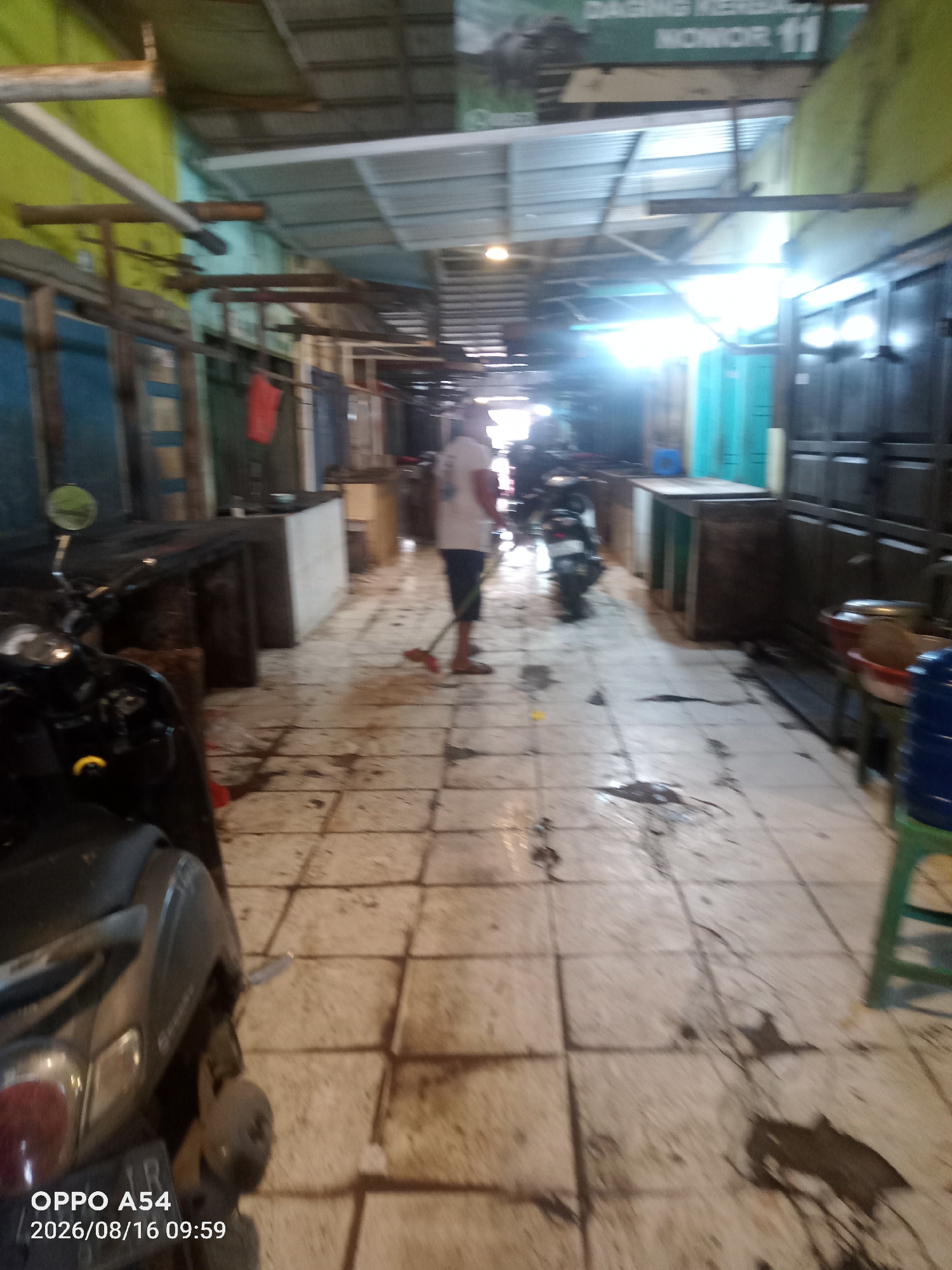 The image depicts an interior view of what appears to be a market or a similar commercial space. The floor is tiled and appears wet and somewhat dirty, with dark splotches and streaks visible. In the foreground, to the left, a dark-colored scooter or motorcycle is parked. A person is visible in the mid-ground, to the right of center, wearing a white t-shirt and dark shorts, holding a broom and sweeping the floor. Several stalls or counters are set up along the sides of the walkway. The lighting is bright in some areas, suggesting artificial light sources, and there are signs visible in the background, though the text is not entirely legible. One sign in the upper background has text in Indonesian that includes "DAGING KER..." and "NOMOR 11". The overall impression is that of a working market, possibly after some activity, with cleaning in progress. The timestamp in the lower-left corner indicates "OPPO A54 2026/08/16 09:59". The location is identified as the city of Jambi, Indonesia.