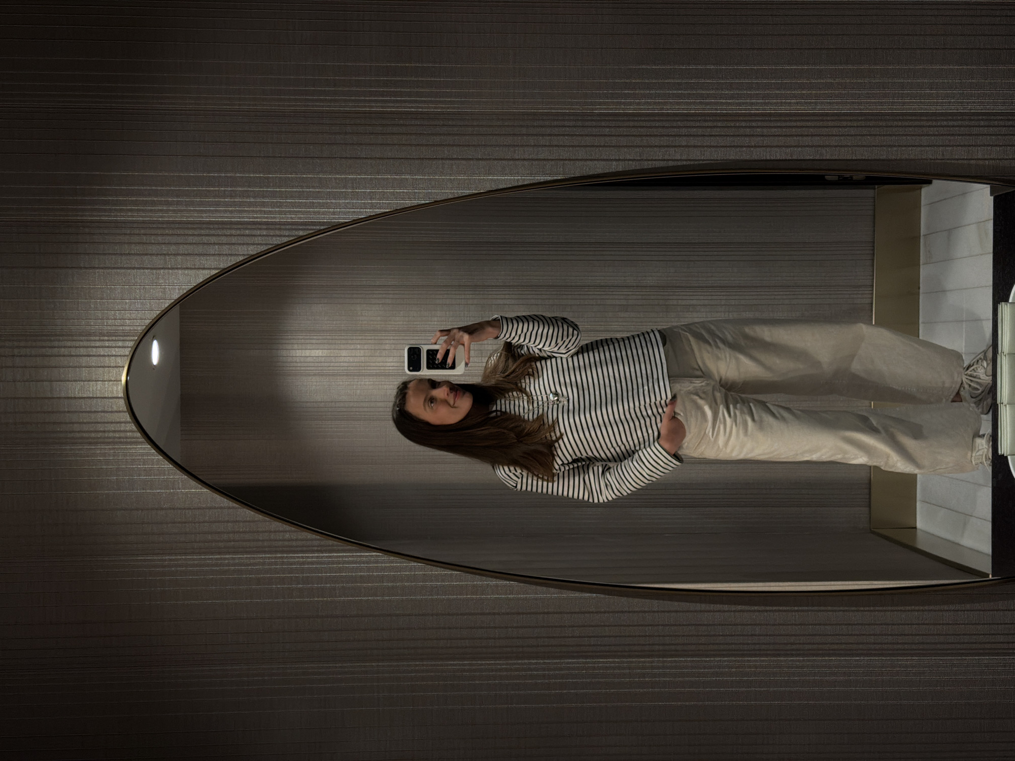 A woman is reflected in an oval mirror. She is holding a cell phone, taking a photo of herself. She is wearing a striped shirt and light-colored pants. The location is Budva, Montenegro. The mirror is mounted on a wall with a horizontal striped pattern. A section of a tiled wall is visible in the lower right corner of the mirror's reflection.