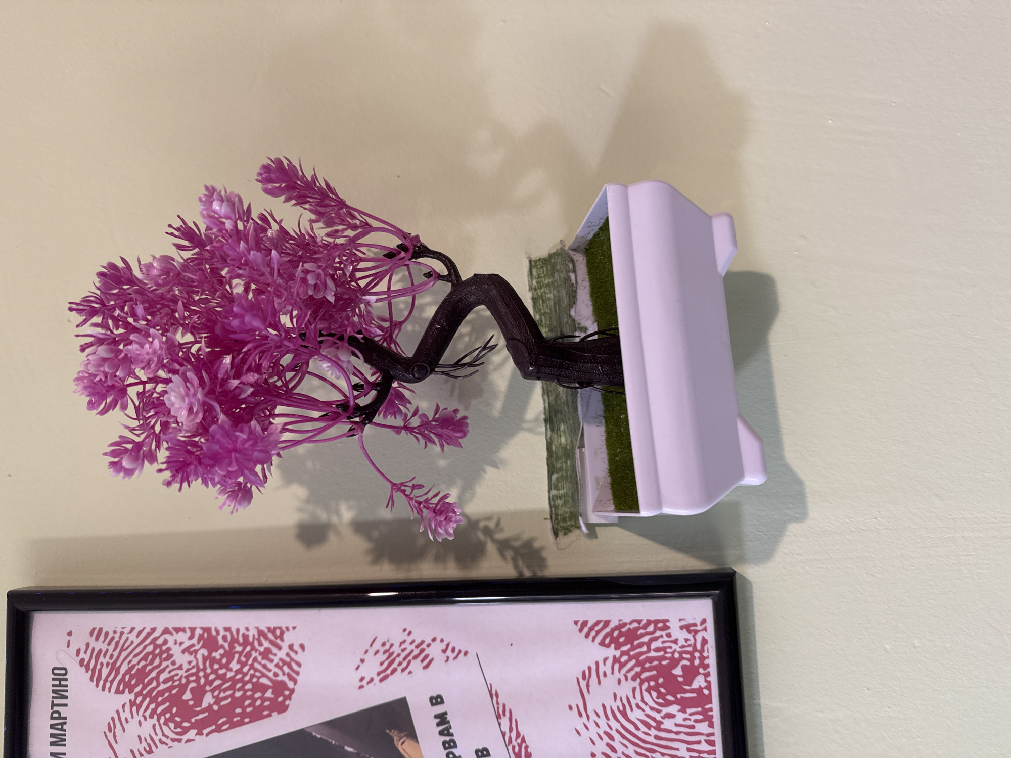 A faux floral arrangement of pink flowers and a dark, synthetic trunk sits in a light lavender-colored container. The arrangement is mounted on a pale yellow wall, positioned above the top edge of a framed poster or picture with a black frame. The poster features red fingerprint patterns and text in a non-English language, possibly Russian based on the letters visible. The scene is indoors. The location in Yana, Bulgaria, is not explicitly identifiable from the visual information.