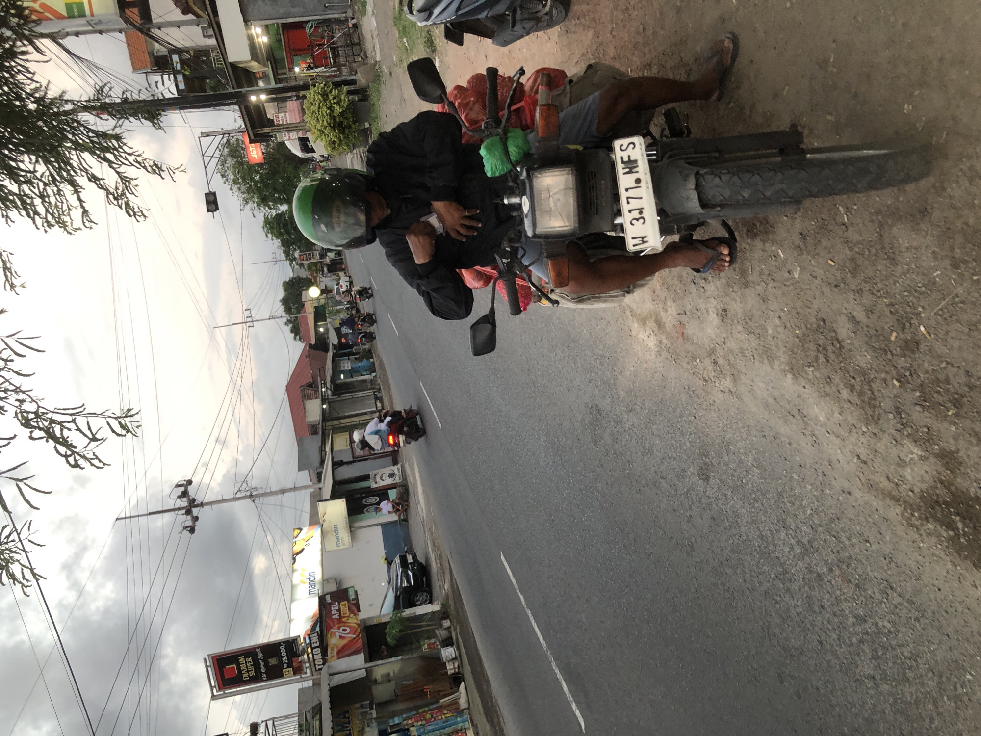 A person wearing a green helmet and a dark jacket is sitting on a motorcycle on the side of a road in Kesambi, Indonesia. The motorcycle is loaded with red bags and green bundles strapped to the back. The rider is reaching into their jacket pocket. The road is paved with white lines and is lined with buildings and shops on the left side of the image. These buildings have signs in Indonesian, including one for "DIARUM SUPER" and another for "mandiri". Power lines are visible overhead. The sky is overcast. Other motorcycles are seen in the background, some with riders.