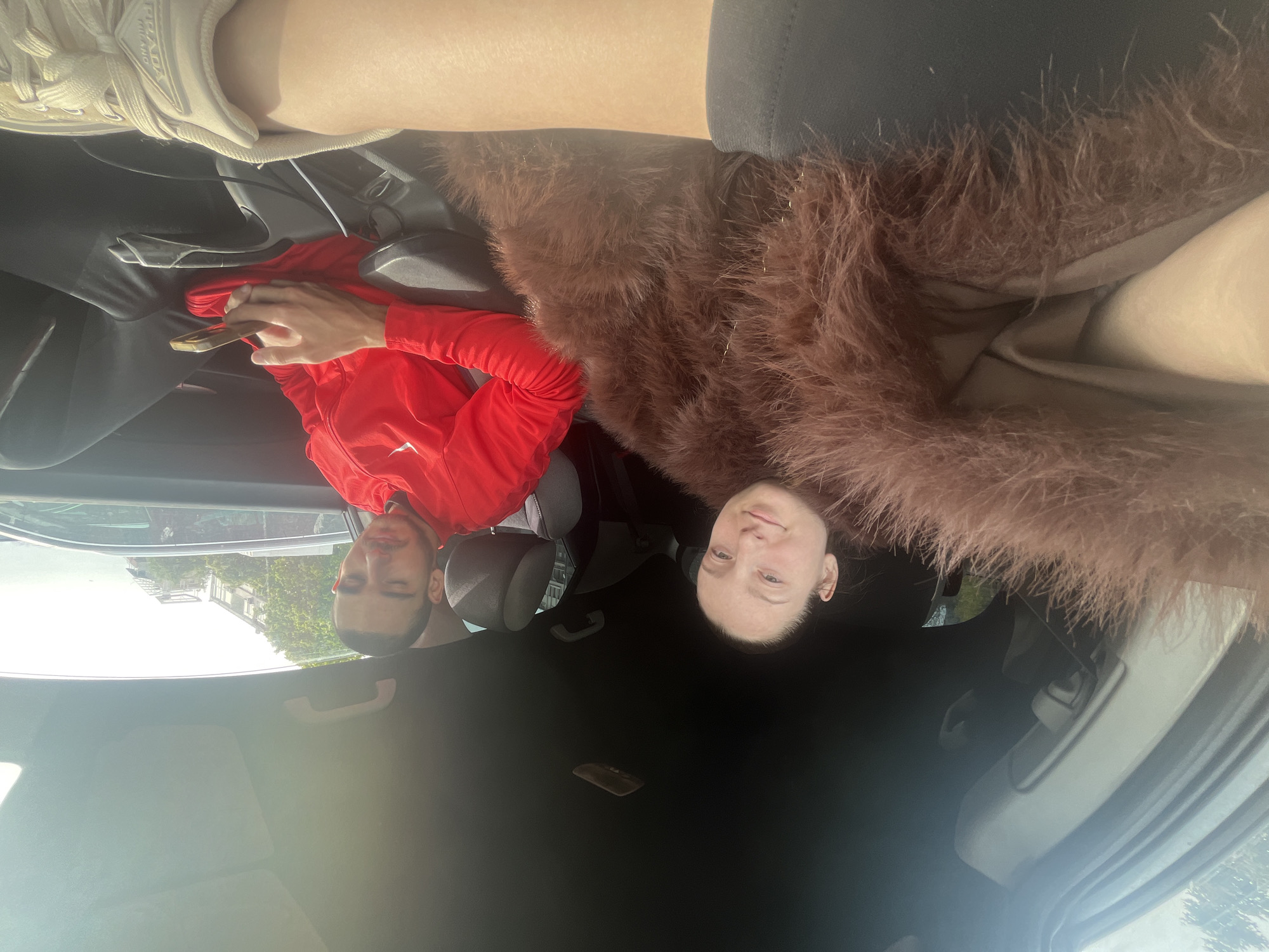 The interior of a car is the primary setting of this photograph. Two people are visible: one individual in a red long-sleeved shirt looking at a mobile phone, and another person with fair skin wearing a brown, textured garment. The car's interior is predominantly black, with seats and dashboard in dark tones. Through the car window, glimpses of buildings and greenery suggest an urban environment. Though it is impossible to say with certainty that this photo was taken in Varna, Bulgaria, there is nothing in the photo that would disprove it.