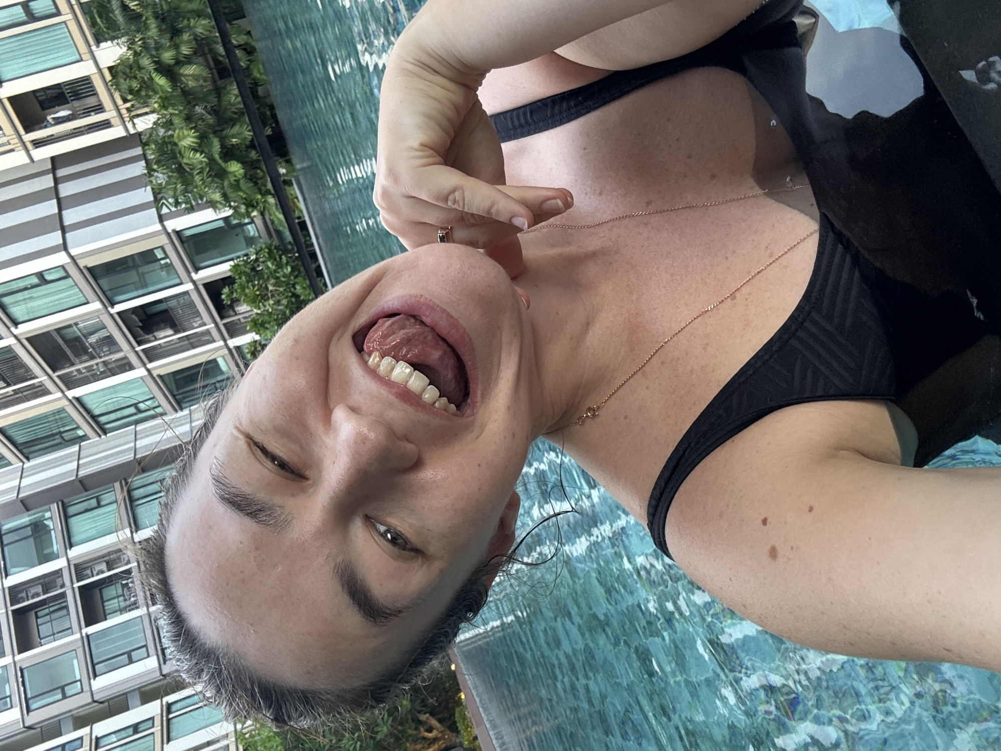 A woman is taking a selfie while partially submerged in a swimming pool. She is wearing a black bikini top and has a playful expression, with her tongue sticking out and her eyes partially closed. Her hand is raised to her face, with a ring on her finger. A thin gold necklace is visible around her neck. The background shows a modern building with many windows and some lush green trees, suggesting an urban or resort setting. The water in the pool is a clear turquoise color with ripples on the surface. The lighting suggests it is daytime, possibly late afternoon given the angle of the shadows. The overall mood is relaxed and joyful.