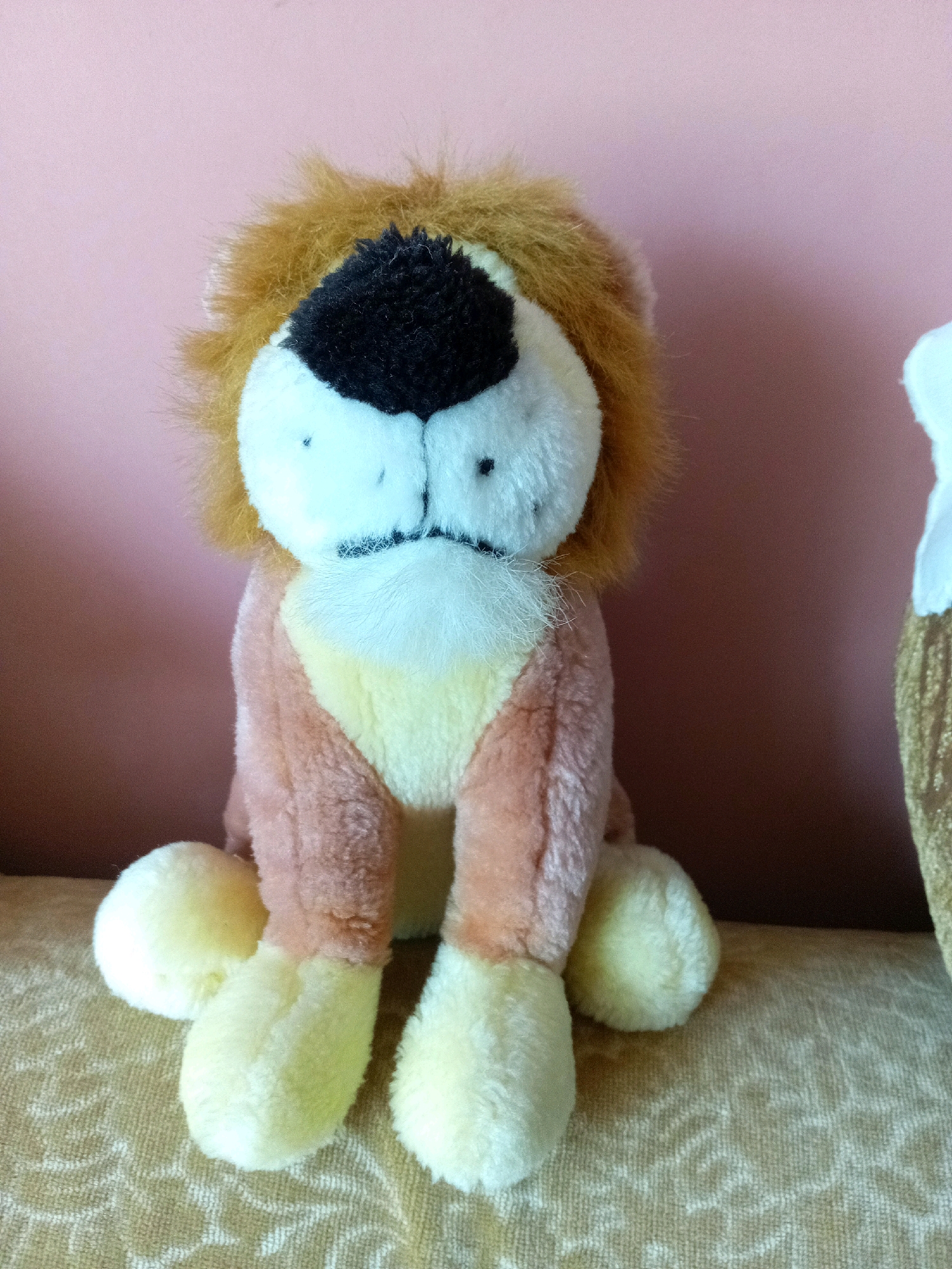 A stuffed lion sits on a patterned surface in front of a pink background. The lion has a mane of brown fur, a black nose, and white fur on its face and paws. Its body is a mix of peach and yellow. To the right of the lion, a portion of another stuffed object, possibly a cone-shaped ornament with gold sequins, is visible. The scene is indoors, and no specific location in Tiachiv, Ukraine, can be identified from the image.