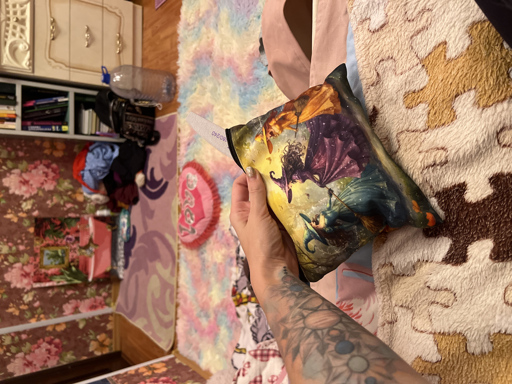 A person's arm with a tattoo is holding a pouch with a fantasy art print of witches. The pouch has a visible label with text. The background is a colorful, soft rug with pastel rainbow colors. To the left, a shelf unit holds books and a bottle of water, a closet with decorative molding, and a floral wallpapered wall. To the right, a textured blanket with a puzzle-like pattern is visible. The scene appears to be indoors, likely a bedroom or living area, with soft lighting suggesting daytime. There are no obvious indications of weather or a specific event, but the overall impression is one of domestic comfort and personal items. The text on the pouch label appears to read "180/240".