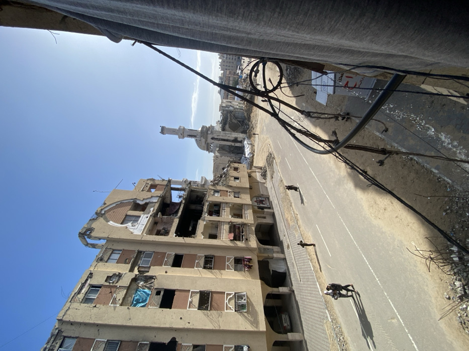 The image depicts a scene of extensive urban devastation under a clear blue sky in Khan Yunis, Palestinian Territory, viewed from a slightly elevated position, possibly a balcony, with a grey fabric and black metal railing partially obstructing the right foreground.
In the midground, an asphalt road with white lane markings runs through the center. On the left side of the road, two individuals walk away from the viewer, one appearing to assist or embrace the other. Further along the left side, a single individual stands facing right. On the right side of the road, another lone individual walks towards the left.
The left side of the image is dominated by a severely damaged multi-story apartment building. It features numerous large structural breaches, exposed interiors, shattered windows, and missing sections of its facade and balconies. A blue tarp is visible in an upper-floor window opening. The ground floor includes arched open spaces, one of which contains a parked white vehicle. Bullet holes or impact marks are visible on the building's exterior.
In the background, positioned centrally, stands a heavily damaged mosque. Its minaret remains partially erect but shows significant structural damage and large cracks. The main body of the mosque is largely reduced to rubble, with skeletal remains of walls and a partial dome visible.
The area surrounding the road and the damaged mosque is strewn with debris and rubble, indicating widespread destruction. In the far background, additional damaged structures and debris fields extend under the bright sunlight. The overall environment conveys a scene of post-conflict desolation with minimal human activity.