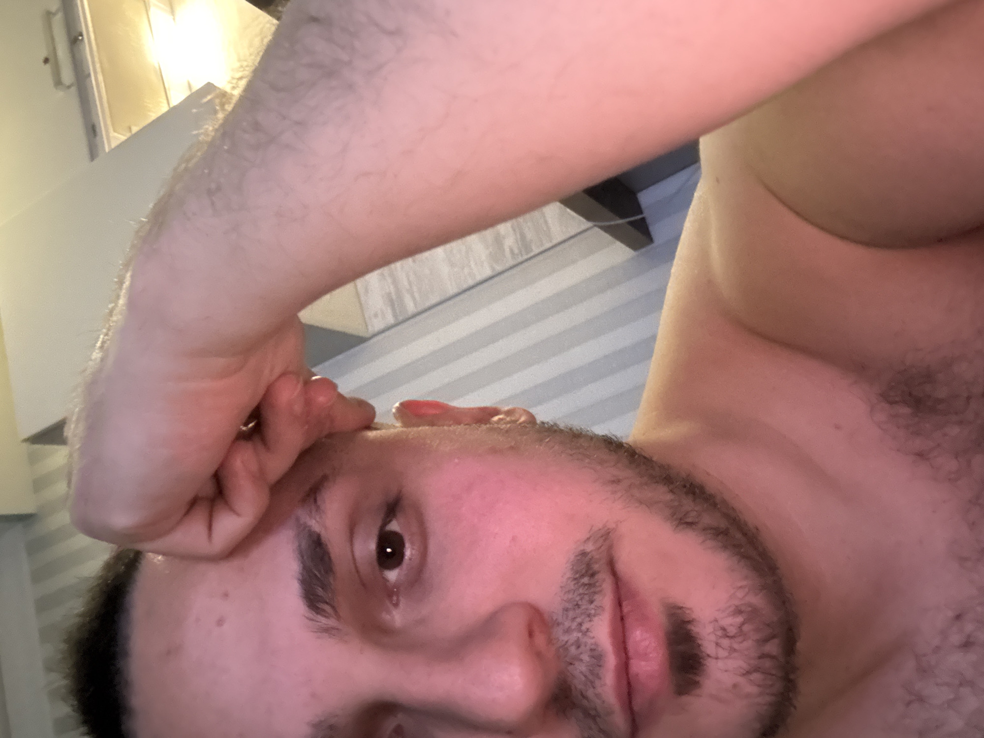 A close-up, angled view of a shirtless man with dark hair and a beard. His right hand is positioned on his forehead. The background includes a wall with horizontal striped paneling and a partial view of a light fixture. The location in the city of Saint Petersburg, Russia is not discernible from the visual data.