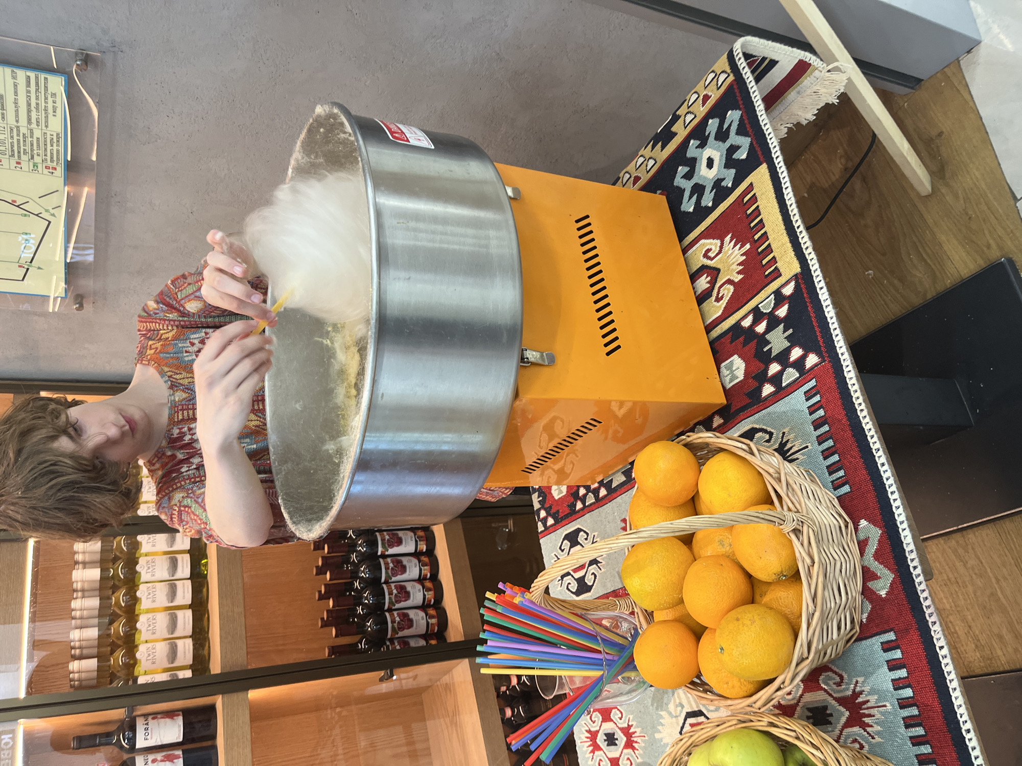 A young person with light brown, wavy hair is making cotton candy in an orange cotton candy machine. The machine is situated on a table covered with a patterned rug. In front of the machine are two baskets of fruit: one filled with oranges and another partially visible with green apples. Behind the machine, shelves are stocked with bottles of wine and juice. The background appears to be a store or a cafe with concrete walls. A map or informational poster is affixed to the wall above the cotton candy machine. The scene is brightly lit, suggesting an indoor setting. The specific location of the city of Perm, Russia, is not discernable from the image itself.