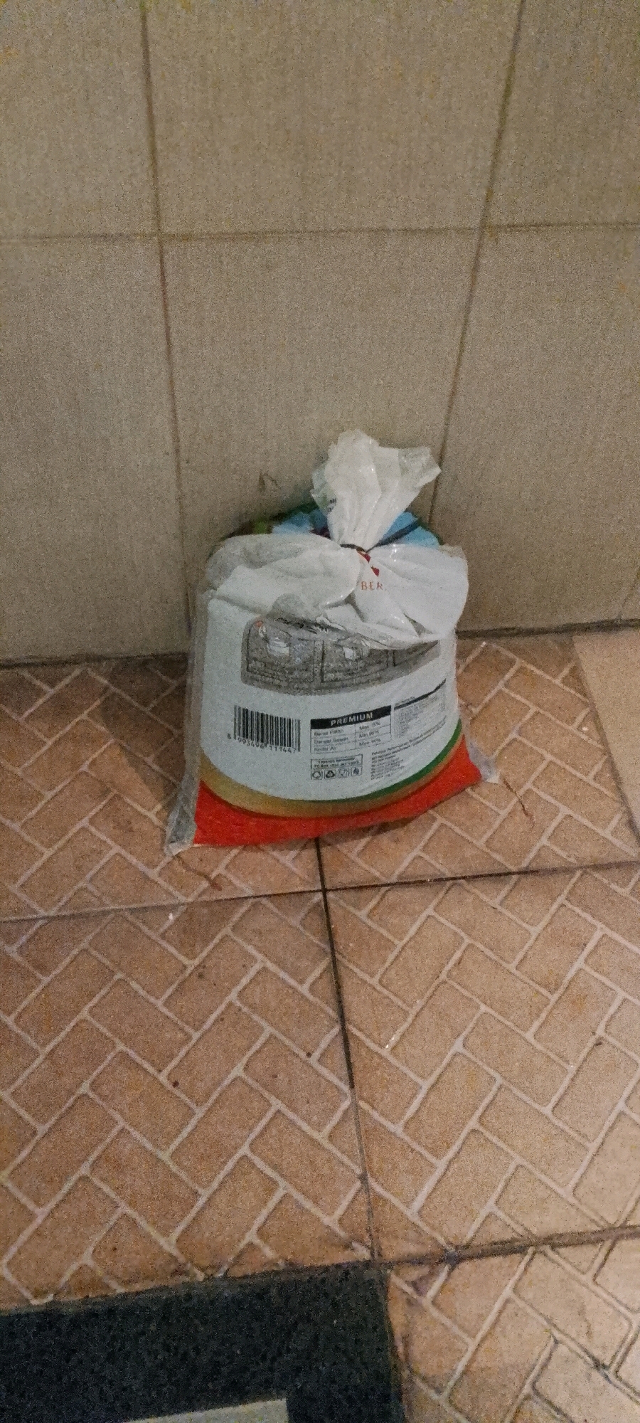 A white plastic bag containing what appears to be pet food is sitting on a tiled floor. The bag is closed with a plastic tie and has some text and graphics on it, including the word "PREMIUM". The floor tiles are a reddish-brown color and arranged in a herringbone pattern. The wall behind the bag is tiled with larger, lighter-colored square tiles. The image appears to have been taken indoors, possibly in a residential or commercial setting in the city of Medan, Indonesia.