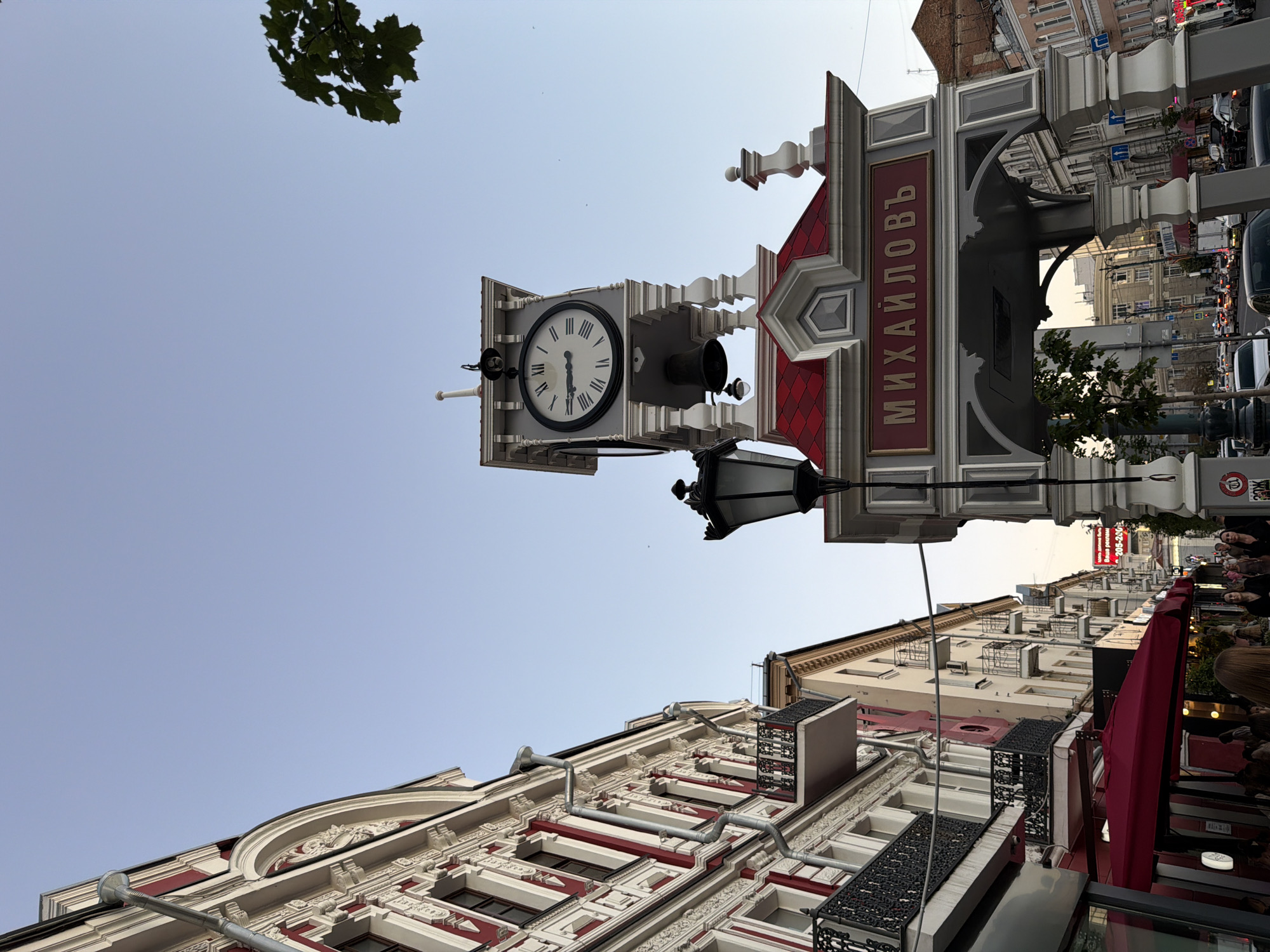 The image shows a street scene in Novaya Usman', Russia. The perspective is from ground level, looking up at buildings and a clock. The orientation of the image is rotated.
The primary subjects are architectural elements of buildings and a street clock. The clock is ornate, with Roman numerals on its face, and is mounted on a decorative post. The post has signage with Cyrillic text that reads "МИХАЙЛОВЪ". Below the clock, a black street lamp is visible. The buildings lining the street display classic architecture with decorative facades, balconies, and red roof accents.
In the background, a street is visible with cars, traffic lights, and more buildings. Some people are present, mostly appearing as blurry figures in the background near storefronts or on the street. One woman is clearly visible in the foreground, on the right side of the image, with her back to the camera.
The time of day appears to be late afternoon or early evening, indicated by the soft, diffused light and the long shadows that would be cast. The sky is a pale blue, suggesting clear weather. The overall atmosphere is that of a bustling urban street with historical architecture.