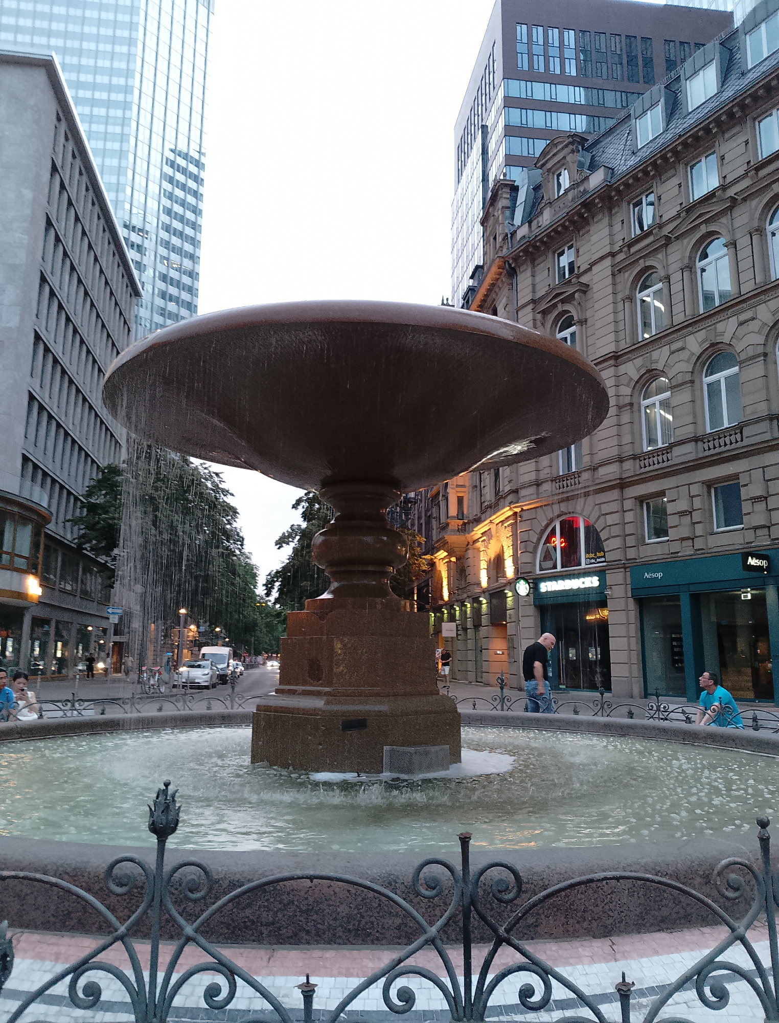 In an urban setting, likely Frankfurt, Germany, a large stone fountain with cascading water is the focal point. The fountain is situated in front of a row of buildings, some modern and tall with glass facades, and others older with ornate architecture and arched windows. A Starbucks is visible on the ground floor of one of the buildings, along with an Aesop store. In the foreground, a decorative metal fence surrounds the fountain. Two men are present near the fountain: one, wearing a black t-shirt and jeans, stands looking down at the fountain, while another, in a blue t-shirt and shorts, sits on the edge of the fountain's basin, facing away from the camera. A street with traffic, including cars and a van, runs behind the fountain, with trees lining the street. The sky is overcast.