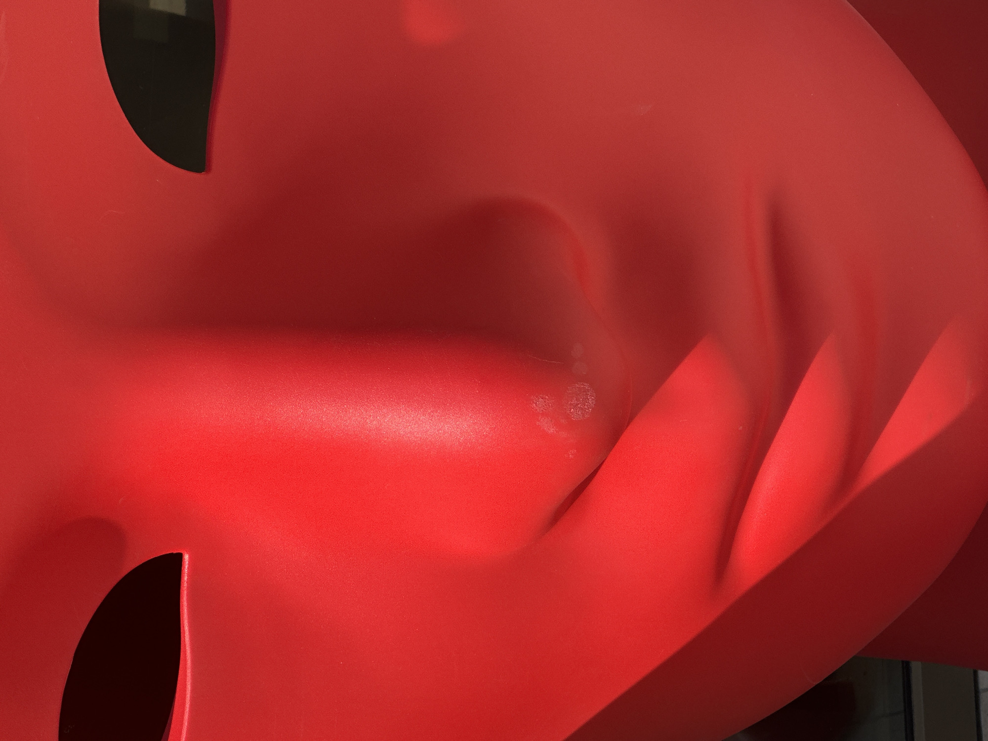 Close-up view of a red, curved, plastic sculpture. The sculpture has smooth surfaces, dark openings, and indented areas. The setting is the city of Budva, Montenegro.