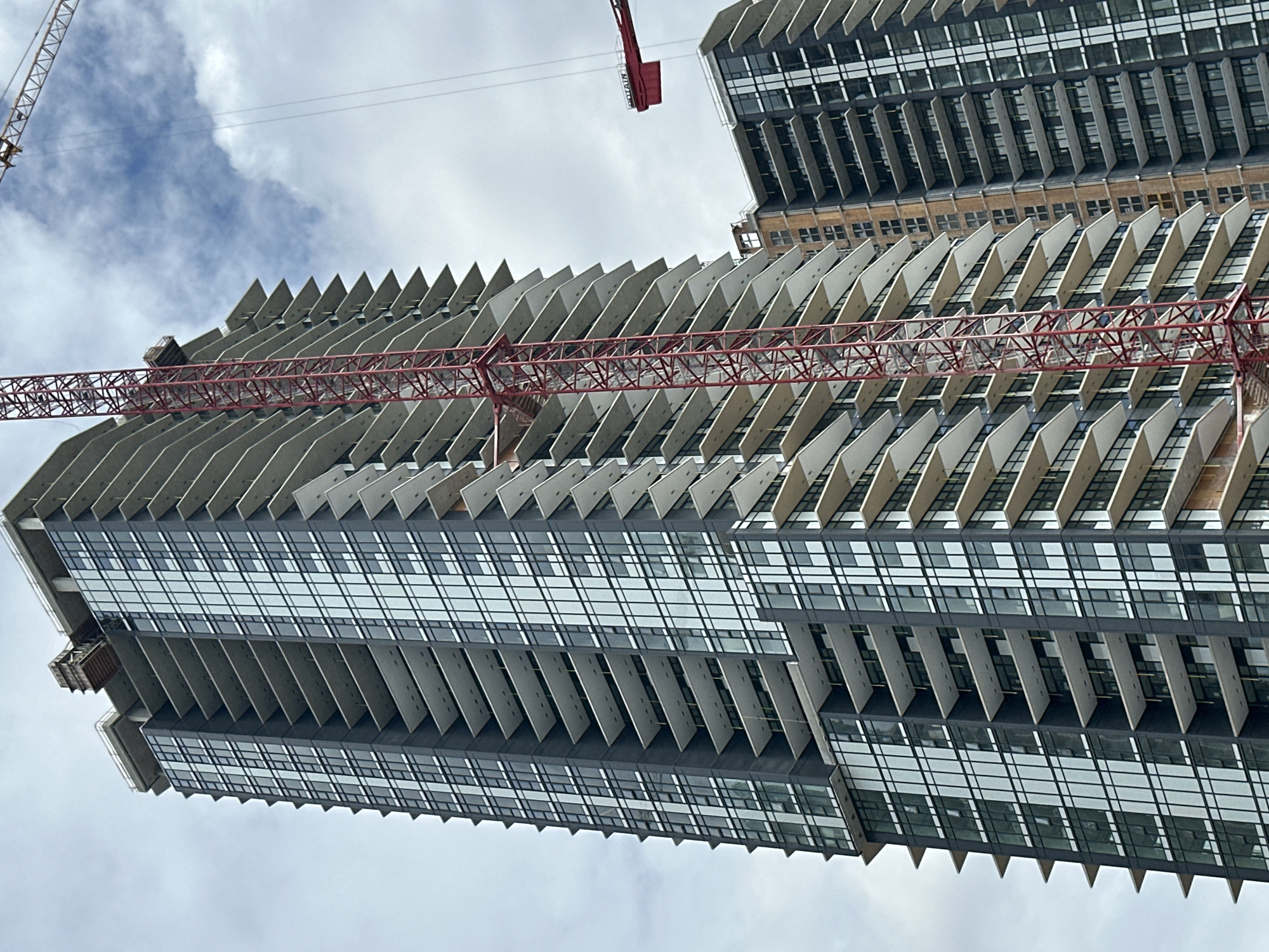 A construction scene in Burnaby, Canada features a partially completed skyscraper with a red construction crane attached to its side. The skyscraper is predominantly clad in dark material and glass windows, with many balconies extending from its facade. Another skyscraper, also under construction, is visible to the right, featuring similar architectural elements. The sky is overcast with some visible clouds. The image appears to be taken from a low angle, looking up at the structures.