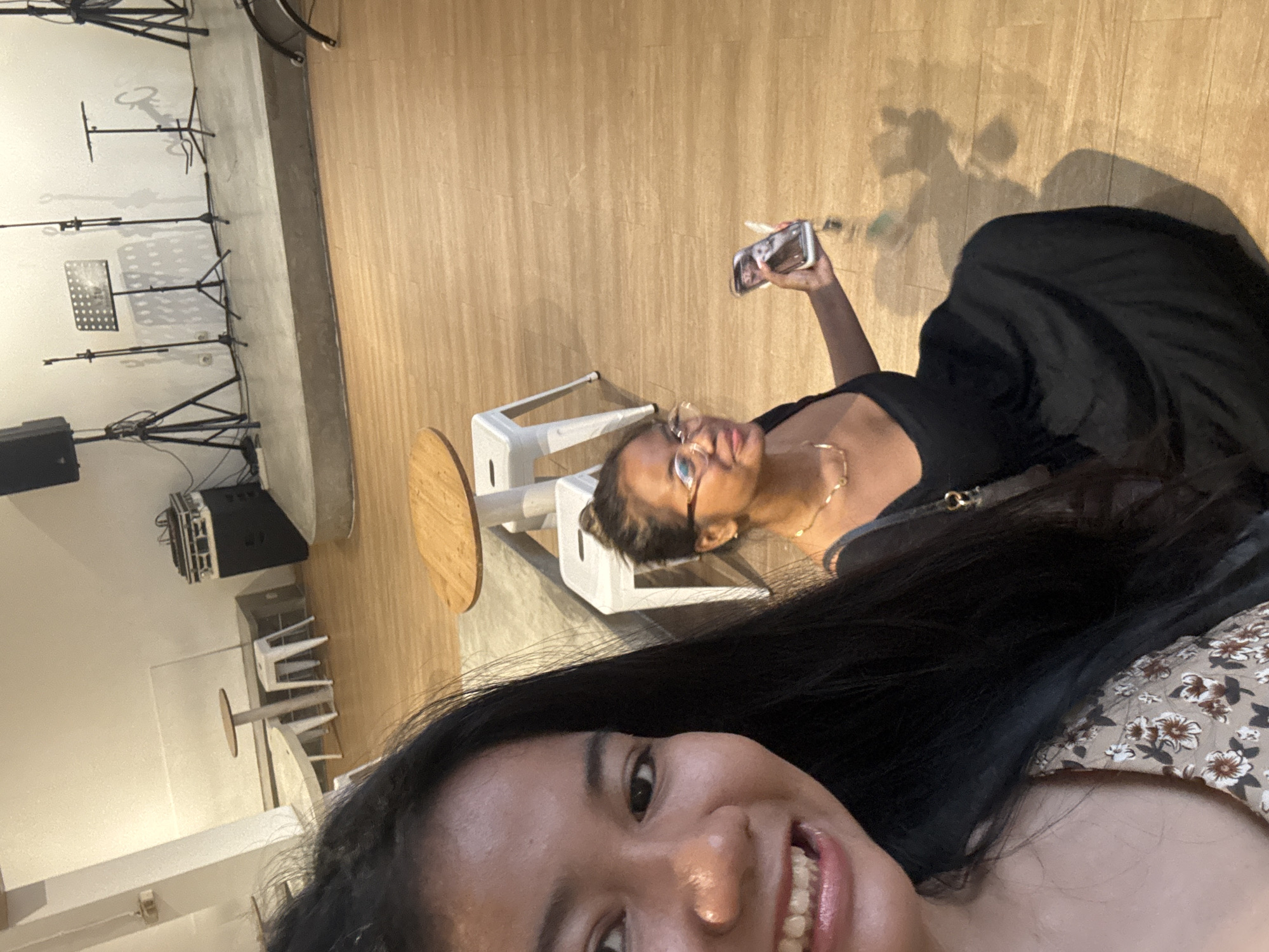 Two women pose for a selfie in a space that appears to be a performance venue or cafe.
One woman, likely taking the photo, is visible in the foreground. She has dark hair and is wearing a floral print top. Only part of her face and shoulder are in view.
The second woman, likely the subject of the photo, is holding a cell phone up to take a photo of herself. She is wearing glasses, a black dress, and a gold necklace. She is partially obscured by a shadow.
Behind them, the space is lit with warm-toned light and shows a wooden floor. Some white chairs and a small round table are visible. In the background, music stands and a speaker are present against a white wall, suggesting a venue for live music.
The location is North Jakarta, Indonesia.