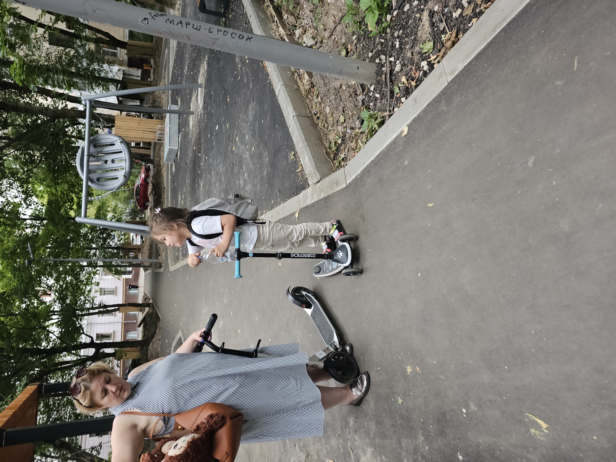 In an outdoor urban park in Khimki, Russia, a woman wearing a striped dress and sunglasses is standing next to a young child. The woman holds a brown purse with a stuffed animal peeking out. The child, wearing a white t-shirt, beige pants, and a backpack, is on a scooter. Another scooter is parked next to the woman. In the background, a playground structure with a circular swing is visible, along with trees and buildings. The ground is paved with asphalt and concrete. A gray pole with Cyrillic writing is on the right side of the image.