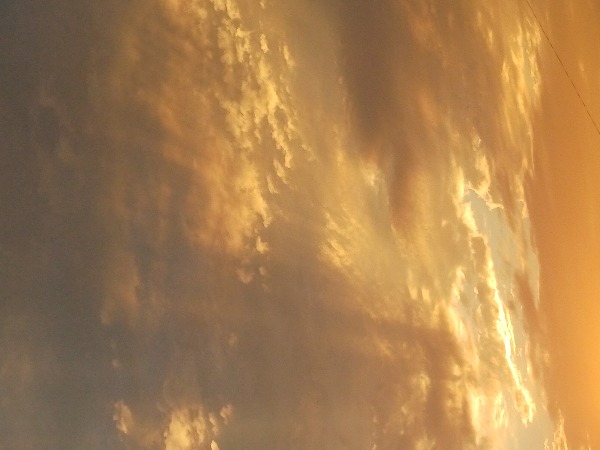 The image is a close-up, vertically oriented shot of a cloudy sky at either sunrise or sunset. The clouds are predominantly in shades of orange, yellow, and gray, with the lower portion of the image showing a brighter, more intense orange hue, suggesting the sun is near the horizon. The lighting creates a dramatic effect, with some clouds illuminated from behind, creating a backlit appearance. The texture of the clouds varies, from wispy and scattered to thicker, more layered formations. There is no discernible urban landscape or specific location within Riga, Latvia visible in the frame. The image solely focuses on the atmospheric conditions of the sky.