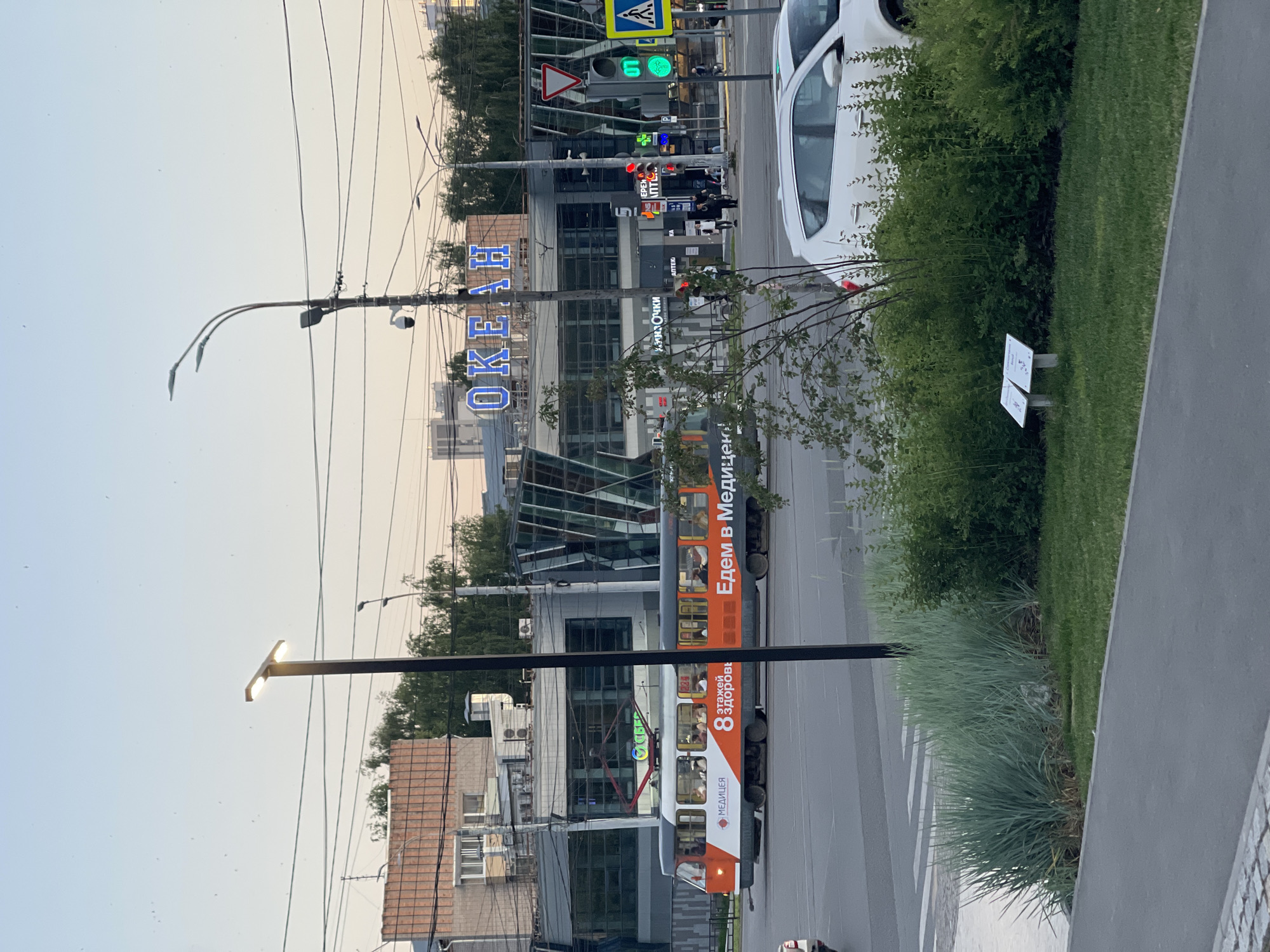 The image depicts an outdoor scene in a city, likely during the late afternoon or early evening based on the lighting. A modern tram is in motion on a paved road, prominently featuring an advertisement for "МЕДИЦЕНТР" (Medical Center) and "ЕДЕМ В МЕДИЦЕНТР" (We are going to the Medical Center). The tram is orange and white and has passengers visible inside. To the left of the tram is a building with a brick facade. In the background, a large modern building with a glass facade displays the word "ОКЕАН" (Ocean) in large blue letters. A traffic light is visible, showing a green light, and a pedestrian crossing signal is also lit. On the right side of the road, a white car is parked or driving. In the foreground, there is a section of green grass and landscaping with various plants, and a paved walkway. There are also power lines and streetlights visible. The overall impression is a busy urban intersection.