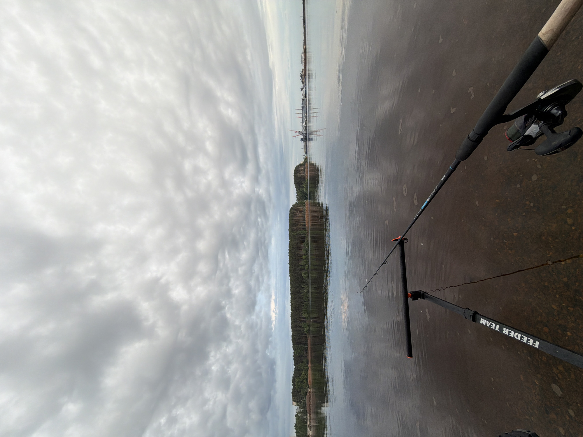 A fishing rod is positioned near the water's edge in Chaikovsky, Russia, with the city's distant buildings visible across the water. The sky is overcast, with clouds dominating the scene. The fishing rod, complete with a reel and line, is set up on a rod rest labeled "FEEDER TEAM." The water reflects the sky and the shoreline, which is lined with trees and vegetation.