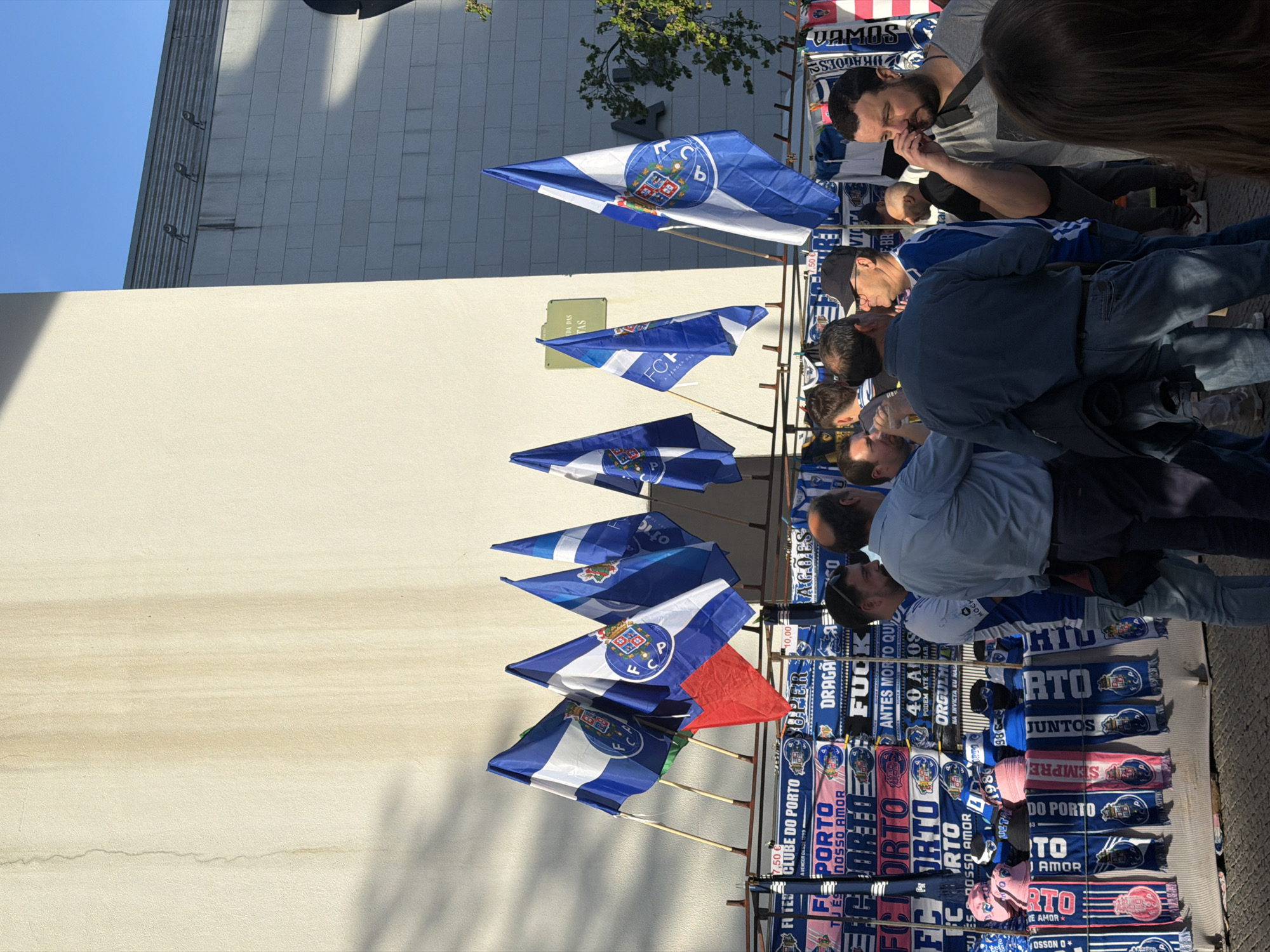The image shows a street scene in Porto, Portugal, where a vendor is selling flags and scarves related to the football club FC Porto. The flags are blue and white with the FC Porto crest. The scarves display club slogans such as "Porto Nosso Amor," "Clube Do Porto," and "Dragão." Several people, presumably fans, are looking at the merchandise. The stall is set up against a plain, light-colored building on a sunny day. There is a sign on the wall behind the stall that has been obscured.