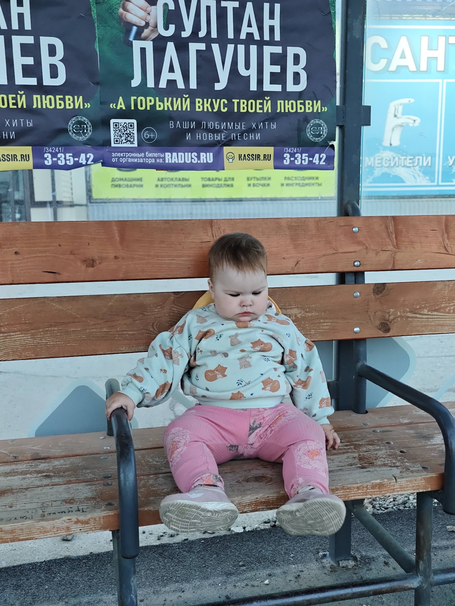 A baby with pink pants, a light blue sweatshirt with animals, and gray sneakers sits on a wooden bench. The bench is located at a bus stop in the city of Chaikovsky, Russia. Behind the bench is a bus stop shelter with posters of Sultan Laguchev with phone numbers on them. Other posters are in the shelter, like a water faucet.
