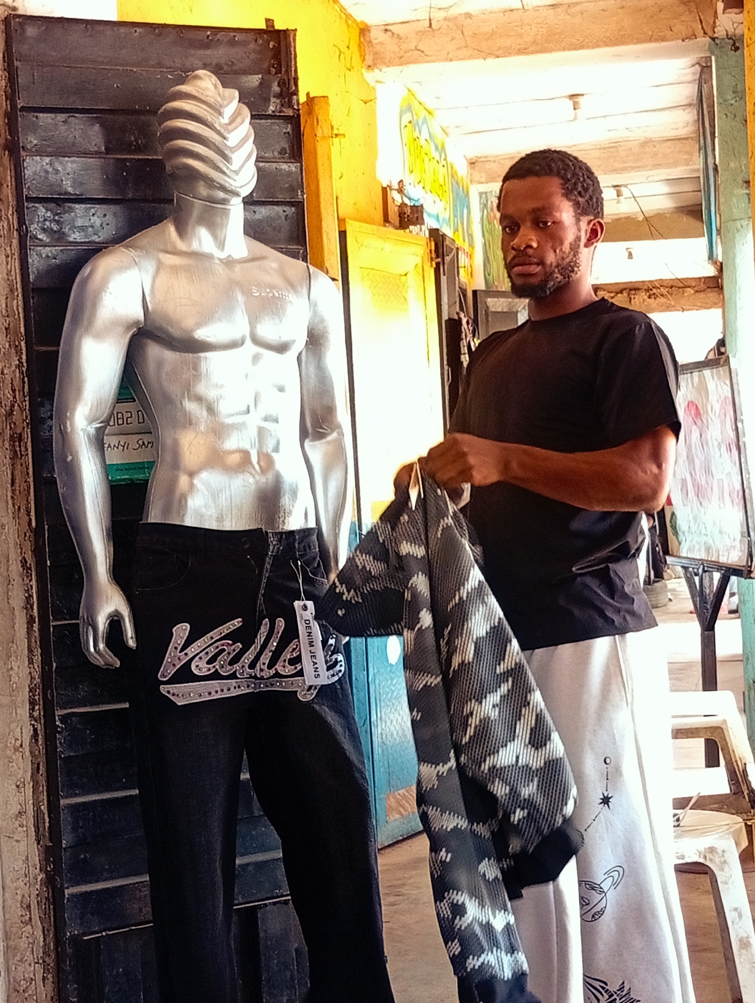 The image depicts a street scene in Bukuru, Nigeria, where a man is standing next to a silver male mannequin. The mannequin is displayed wearing dark jeans with a prominent "Valley" logo adorned with rhinestones and a "DENIM JEANS" tag. The man, wearing a black t-shirt and light-colored pants with illustrations of planets and stars, is holding a dark patterned garment.
The setting appears to be an outdoor market or shopfront, characterized by a vibrant yellow wall, dark wooden slatted doors, and visible ceiling structures with exposed wiring and lights. The overall atmosphere suggests daytime, with bright, natural lighting illuminating the scene. There are also white plastic chairs visible in the background, hinting at a commercial or public space.
The expression on the man's face is neutral, and his interaction with the clothing suggests he might be a vendor or a customer. The background elements, including the graffiti-like artwork on the wall and the rustic appearance of the shopfront, provide strong cues about the location being an urban area in Nigeria. The image is well-lit, showcasing the details of the clothing and the surrounding environment.