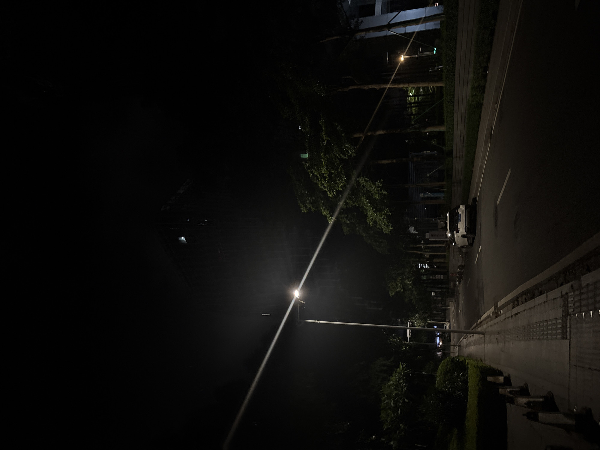 This image depicts a nighttime street scene in Haizhu District, China. The composition is dominated by darkness, with artificial lighting providing the primary illumination. A single, bright street lamp casts a long beam of light diagonally across the frame, cutting through the gloom.
To the right of the street lamp, a paved sidewalk is visible, bordered by neatly trimmed bushes and trees. Beyond the sidewalk, a road stretches into the distance, reflecting the ambient light. A white vehicle, possibly a car or van, is parked along the side of the road. In the background, the facade of a modern building is partially illuminated, suggesting an urban setting. Trees are scattered throughout the scene, their leaves catching the light in places.
The time of day is clearly night, indicated by the darkness and the artificial lighting. The weather appears clear, as there are no visible signs of rain or fog. The scene evokes a sense of quiet solitude and urban stillness, typical of a street at night. There is no visible text in the image. The overall impression is one of a peaceful, albeit dark, urban environment.
