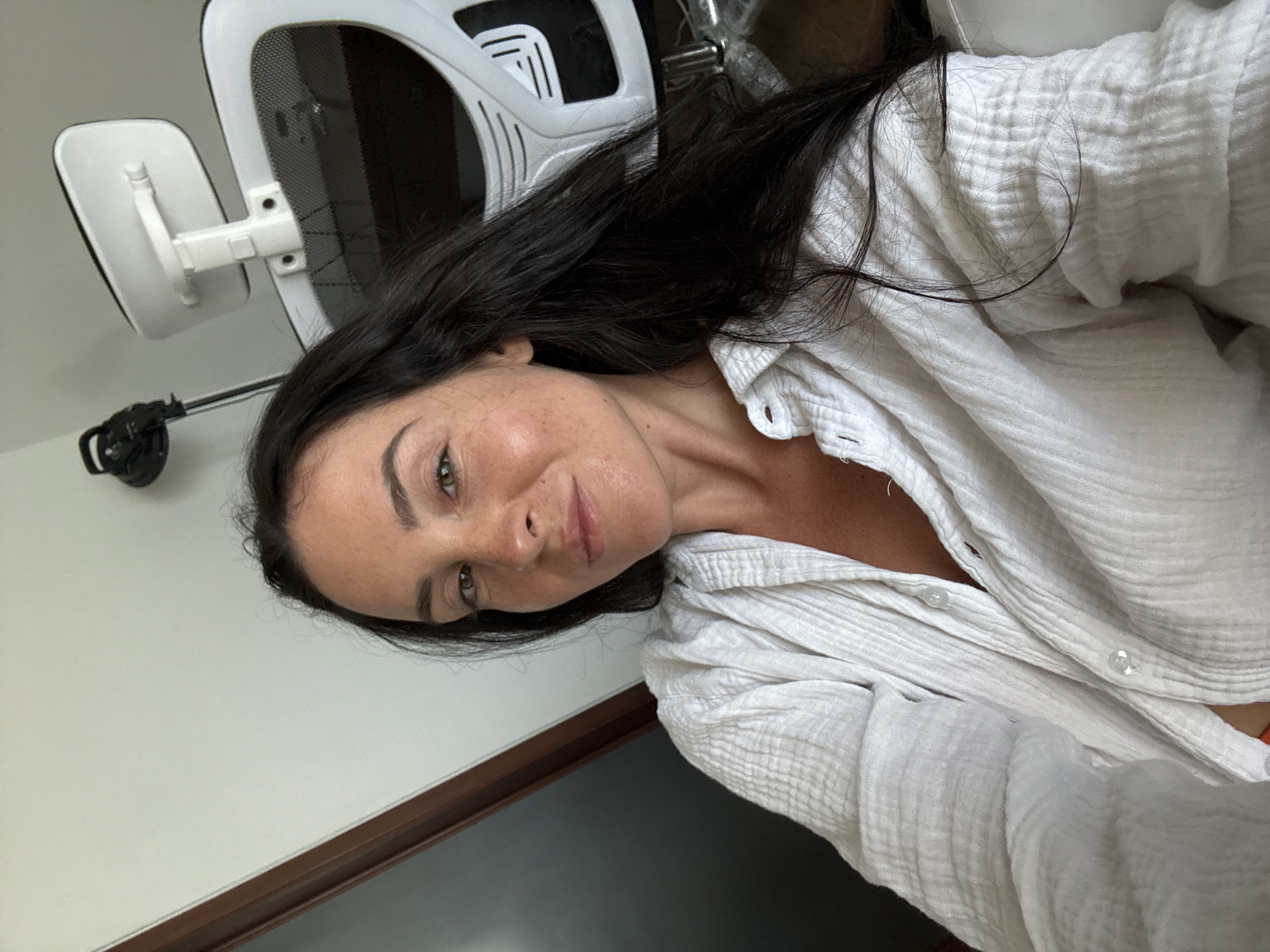 A selfie taken from a low angle shows a woman with long, dark hair and brown eyes. She is wearing a white, textured button-down shirt. Behind her, a white office chair with a mesh back is visible. The background consists of a light-colored wall and a dark wooden doorframe. The scene appears to be indoors, possibly in an office or home setting in North Kuta, Indonesia.