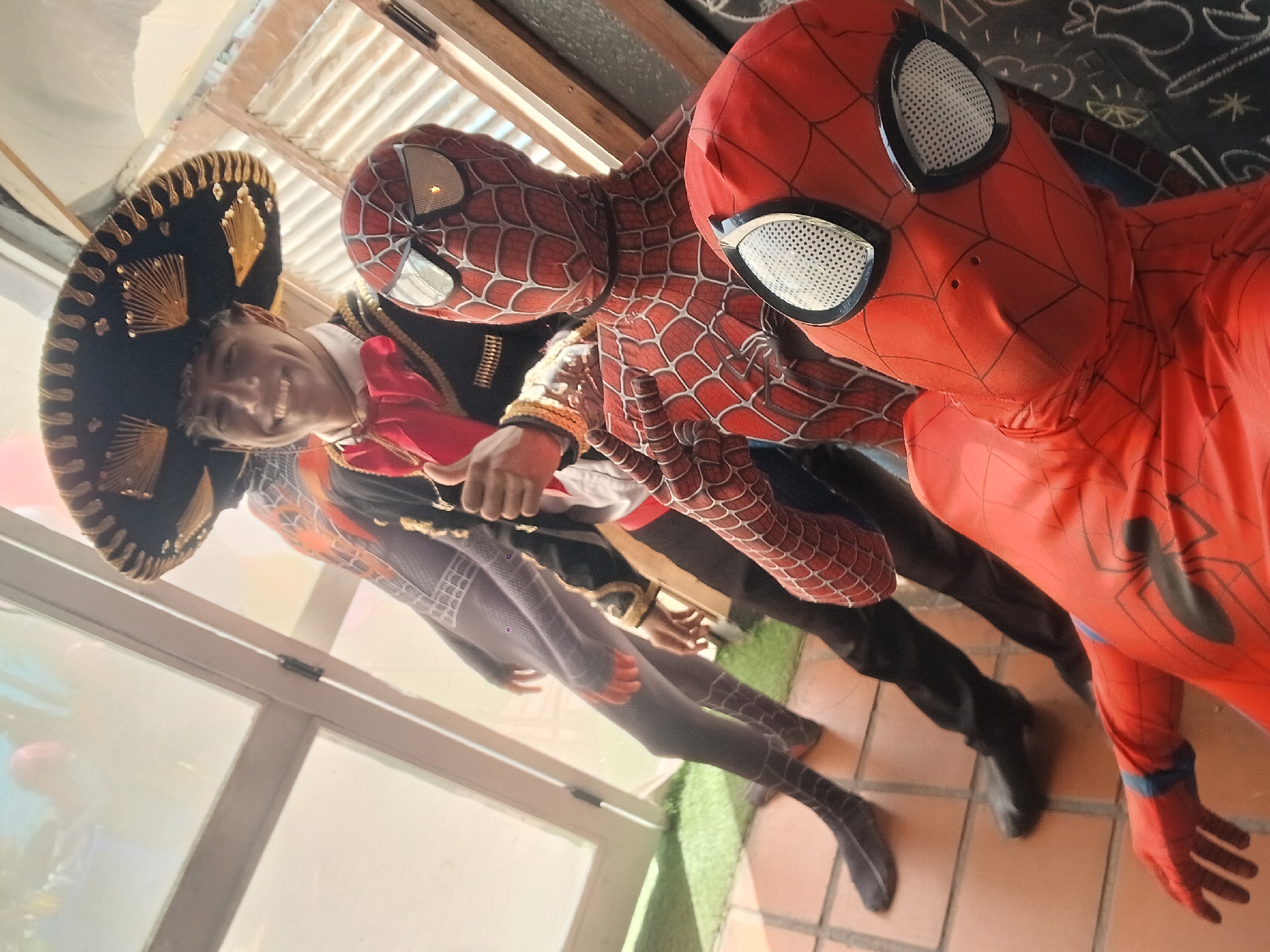 Three people are posing for a picture. Two are wearing Spider-Man costumes. One wears a traditional mariachi costume with a black sombrero. They are on a tile floor in what looks like a restaurant or business establishment. This image was taken in Porlamar, Venezuela.