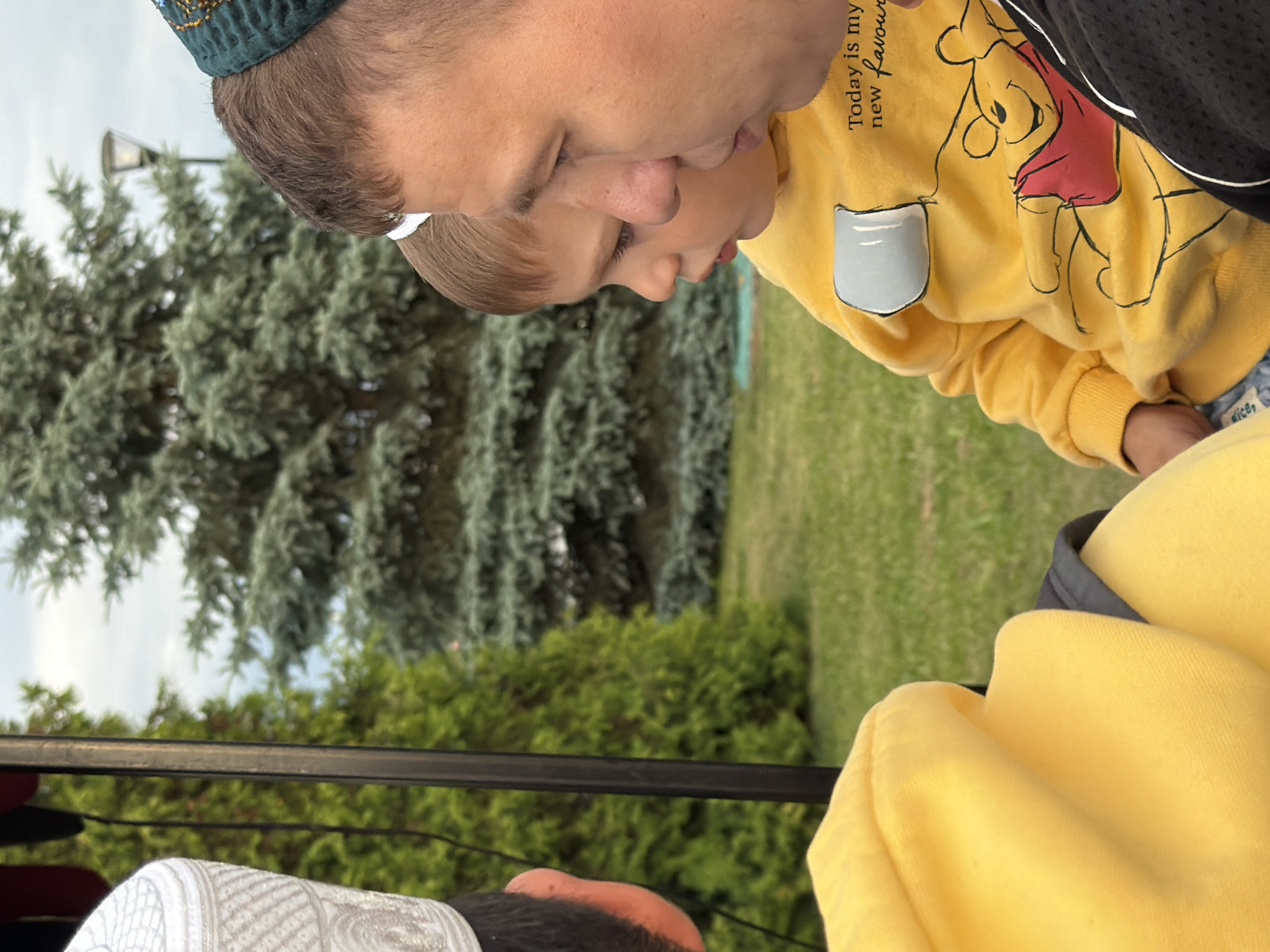 A close-up, side-profile shot shows an adult man and a young child looking towards the left of the frame, with trees and a lamppost in the background. The adult wears a green, embroidered cap. The child is wearing a yellow sweatshirt with a Winnie the Pooh design and the text "Today is my new favourite". The adult's right shoulder is visible on the right side of the frame. Another person's left shoulder, wearing a white cap, is visible on the far left. The scene is outdoors, with green grass and trees.