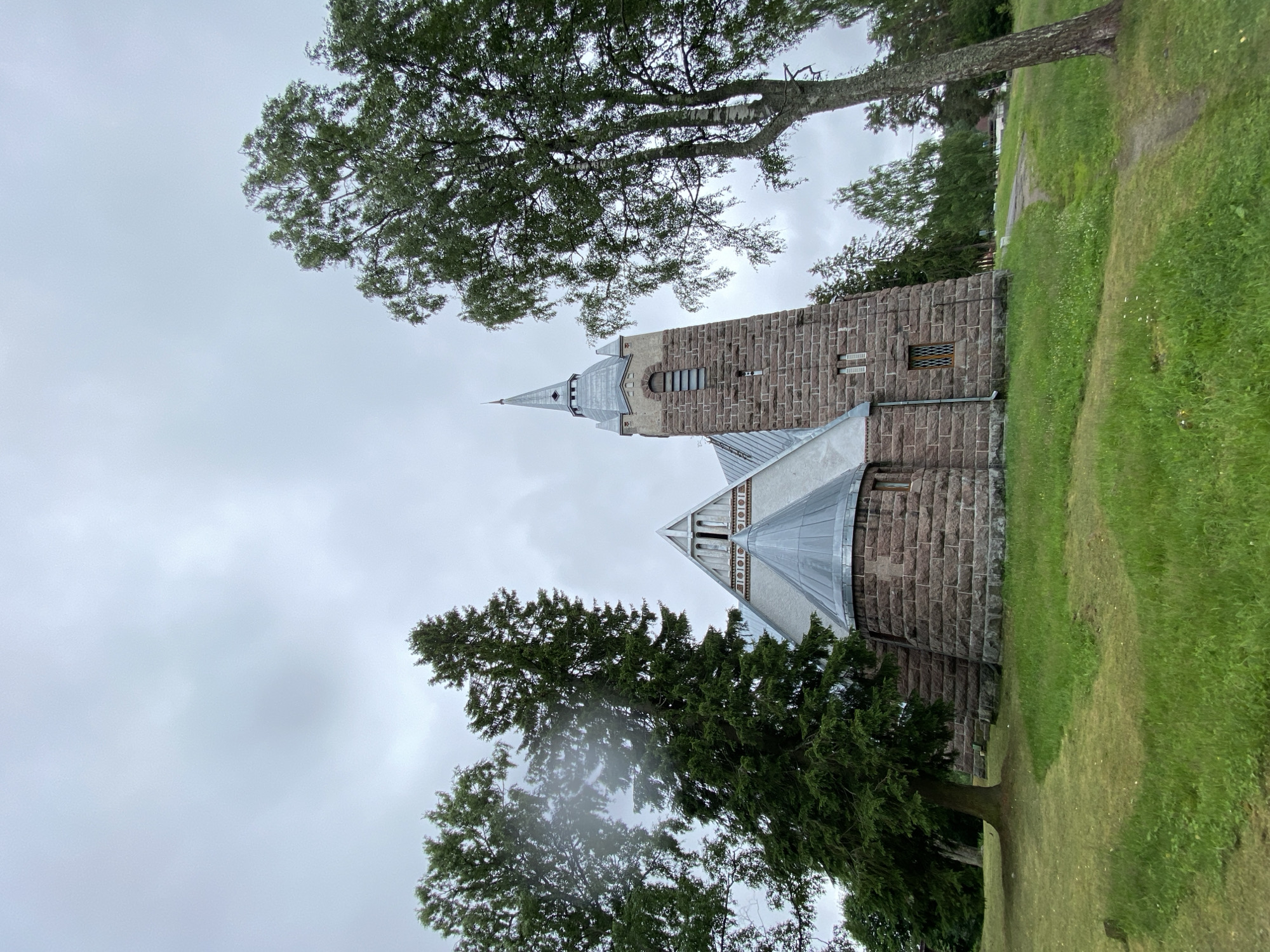 An exterior, low-angle, eye-level shot captures a stone church with a steeple and a peaked roof, set against a cloudy, overcast sky. The church's facade is constructed from rough-hewn, reddish-brown stones. A tall, dark green evergreen tree is positioned to the left of the church, and a lighter-barked tree with green foliage stands to the right, partially obscuring the view of the church's right side. A path winds through a grassy area in front of the church, which slopes upwards towards the building. The scene is located in the city of Melnikovo, Russia.