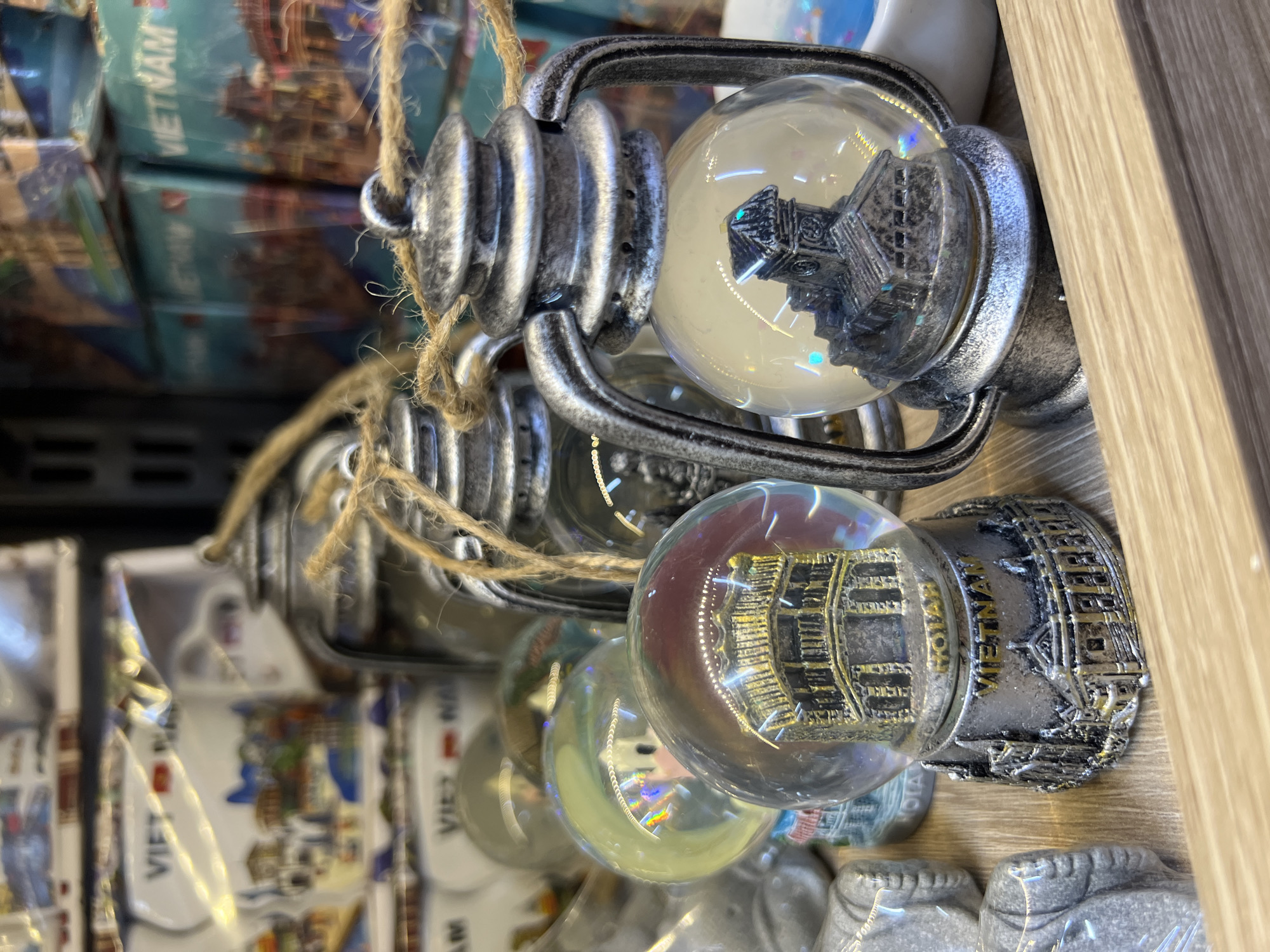 The image displays a close-up, shallow depth-of-field view of souvenir items for sale. In the foreground, two miniature, decorative lanterns with glass spheres are prominently featured. Each lantern has a silver-toned, antique finish and is adorned with twine. Inside the glass spheres are miniature architectural models, possibly of historical buildings or landmarks. One sphere contains a detailed replica of a multi-story building with arched windows, sitting on a base that reads "VIETNAM". The other sphere, to the left, contains a similar architectural structure with a golden hue and is also on a base. The bases of both items are intricately designed with architectural motifs and text. In the background, other souvenirs are visible, including what appear to be snow globes and stacked boxes with "VIETNAM" printed on them. The setting appears to be a retail environment, possibly a souvenir shop. There are no people or animals present in the image, and no specific location within Nha Trang, Vietnam, can be identified from the visual information provided. The lighting is soft and diffused, highlighting the details of the souvenirs.