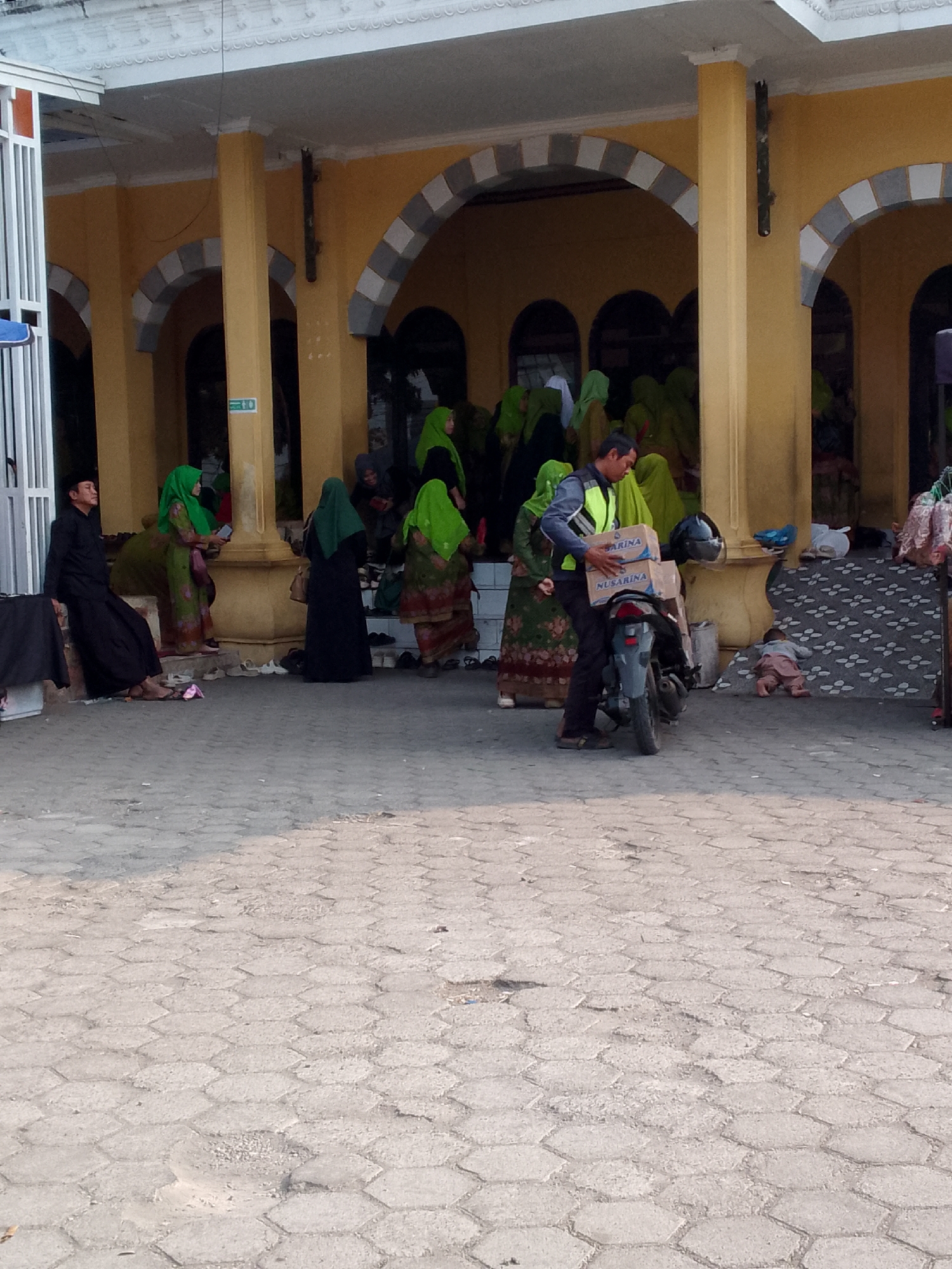 The image depicts an outdoor scene in front of a building with yellow walls and arched openings. Several people are present, most of whom are wearing traditional Indonesian attire, including headscarves and long dresses. In the foreground, a man in a reflective vest is standing next to a motorcycle, holding two cardboard boxes. A baby is lying on the ground near the motorcycle. The ground is paved with hexagonal interlocking concrete blocks. The overall impression is of a public space, possibly near a religious site given the attire of the people. The location is identified as Rajamandala Kulon, Indonesia.