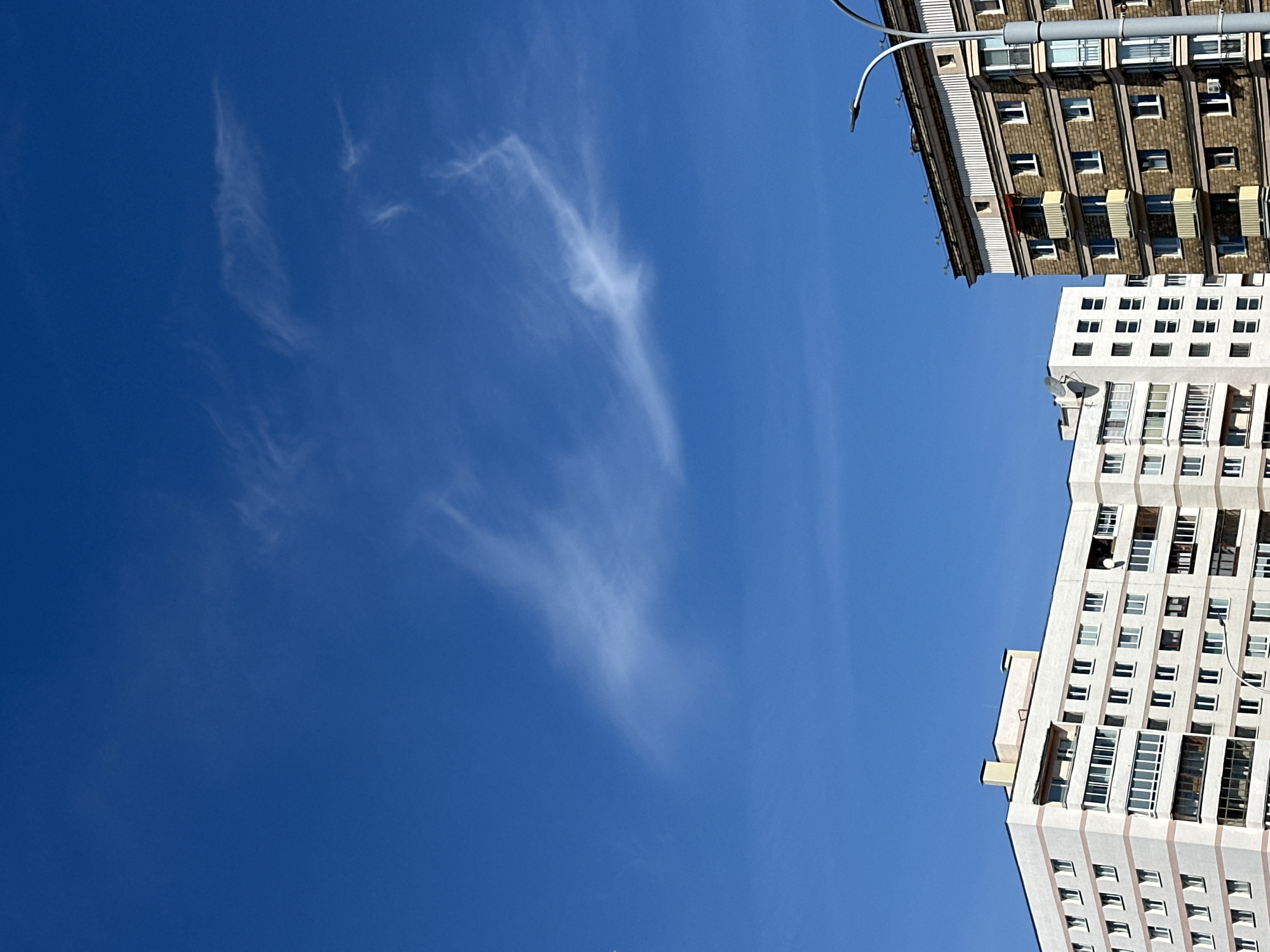 A vertical, low-angle shot shows the bright blue sky above two apartment buildings. The building on the left is white with a few pink bands and numerous windows, some of which are enclosed balconies. The building on the right is made of brown brick with white trim and balconies with yellow awnings. A streetlamp on the right side of the frame arches over the top of the brick building. Wispy white clouds are scattered across the sky. The buildings appear to be in a residential area of Minsk, Belarus, given the architecture typical of post-Soviet urban planning. No people are visible in the image.