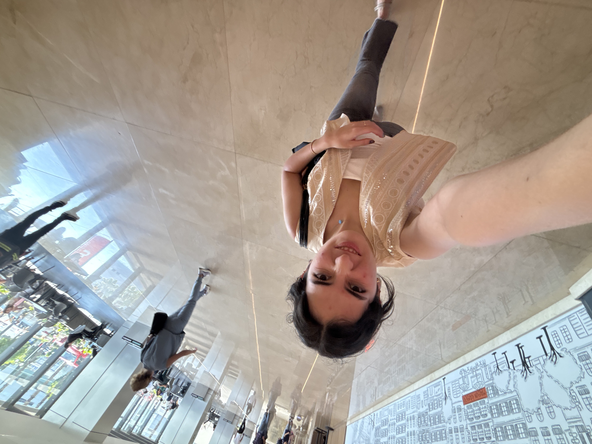 A woman is taking a selfie in what appears to be a commercial or government building in the city of Puebla, Mexico. The floor is shiny tile, and there is a mural on the wall. The woman is wearing jeans, a white top, and a sequined vest. She has dark hair and is looking at the camera. The building's windows reflect the outdoors and people on the street outside. The reflection is of a street scene from the outside including a building with a red sign.