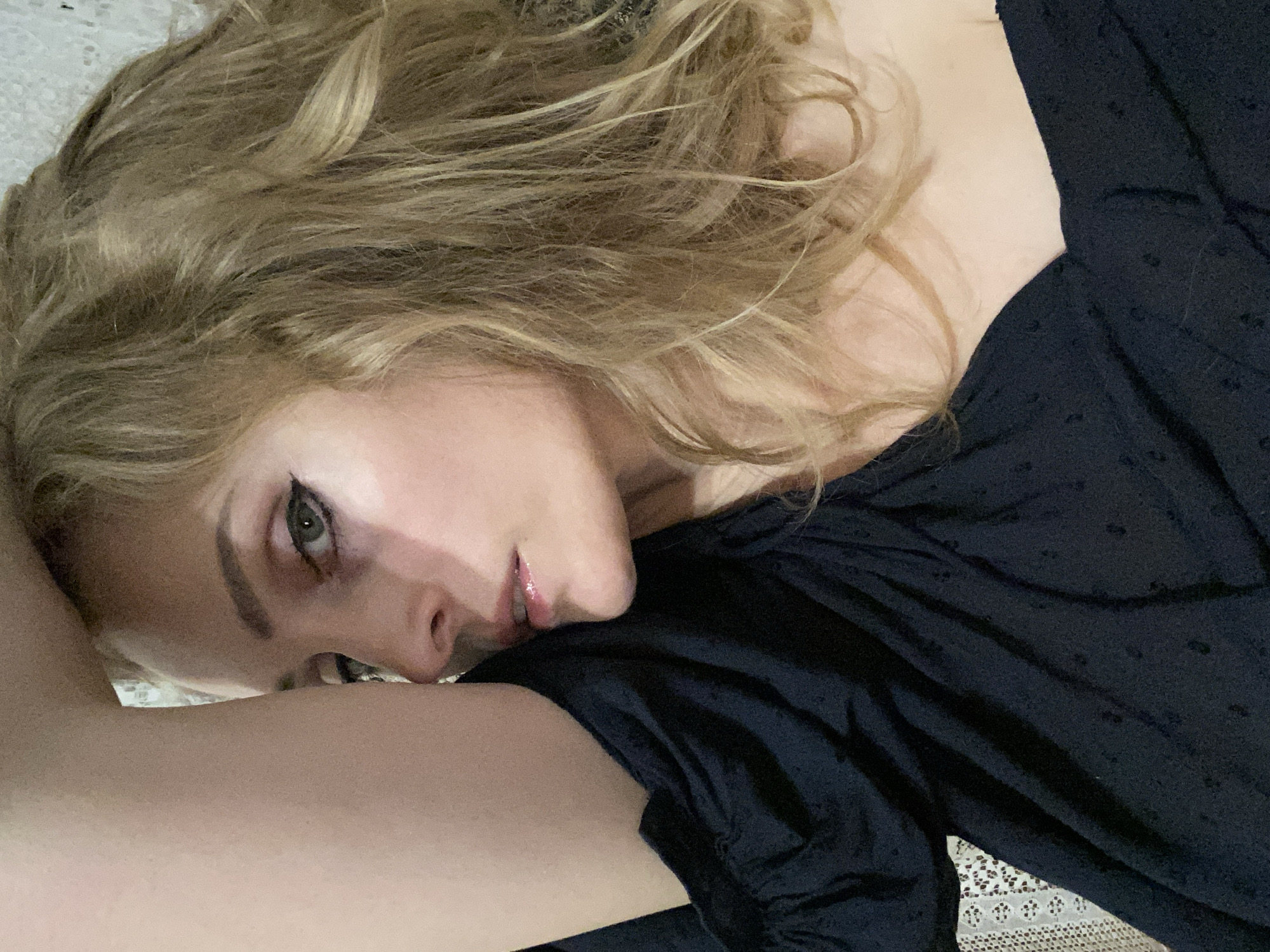 The image is a close-up portrait of a woman with blonde, wavy hair. She is wearing a dark, off-the-shoulder top with a subtle pattern. Her left arm is raised and bent, obscuring part of her face. She has green eyes and is wearing eyeliner. Her lips are slightly parted and appear to be glossy. The background is softly blurred and appears to be a textured surface, possibly a wall or fabric. There is no indication of a specific location within Lida, Belarus, in this image.