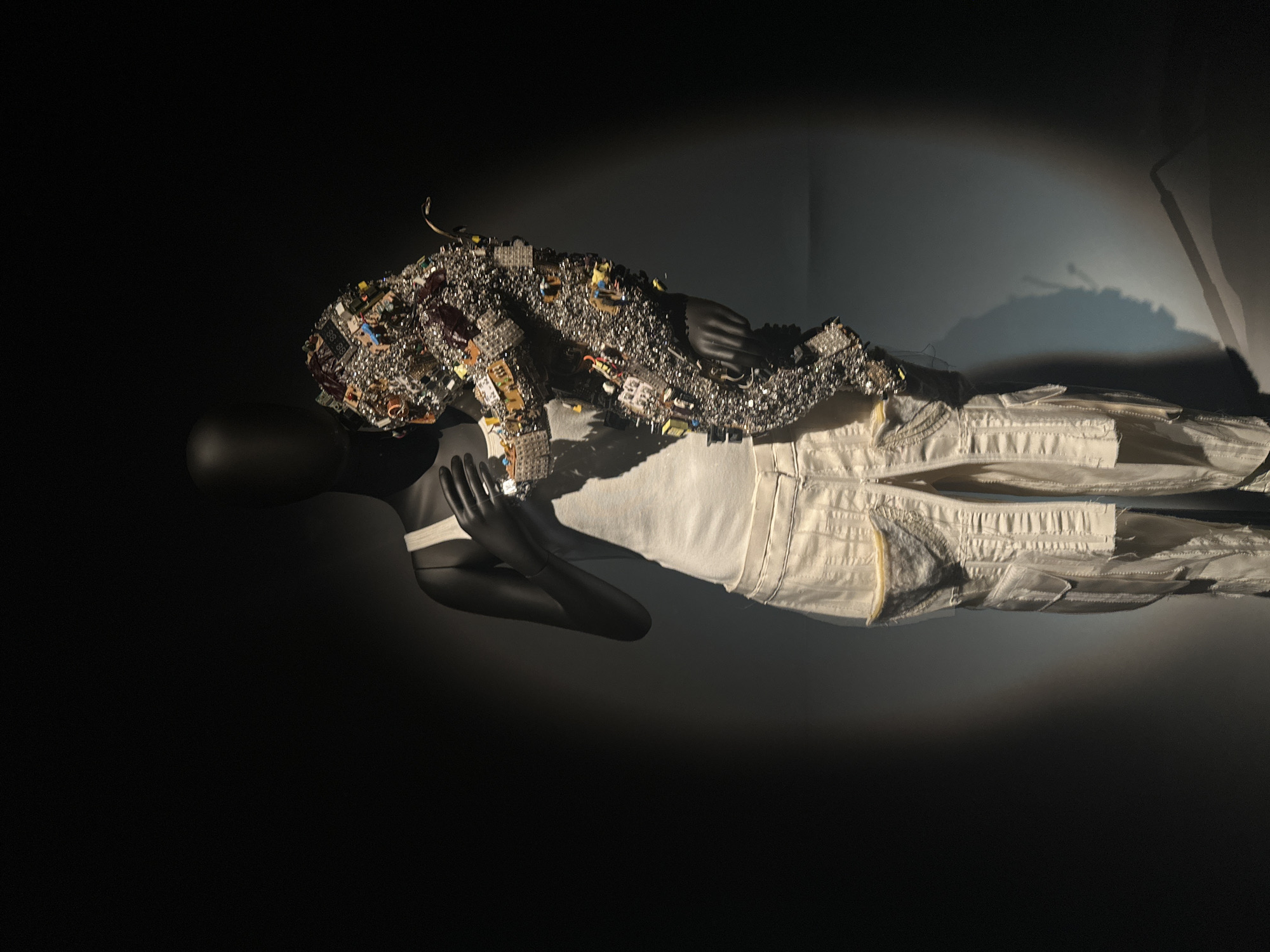 A full-body, dark grey, headless mannequin stands upright against a dark background, illuminated by a directed spotlight from the upper left. The mannequin is dressed in a plain white sleeveless tank top and light beige cargo trousers with multiple pockets and visible stitching. Its arms are positioned to cradle an abstract, organic-shaped sculpture resembling a stylized animal, possibly a fox, against its left side. This sculpture is intricately constructed from numerous small electronic components, including circuit board fragments, resistors, capacitors, and metallic pieces, creating a complex, textured surface primarily in silver and dark grey with various colored accents. The exhibit is located within the Design Museum at 224-238 Kensington High Street, Kensington, London, United Kingdom.