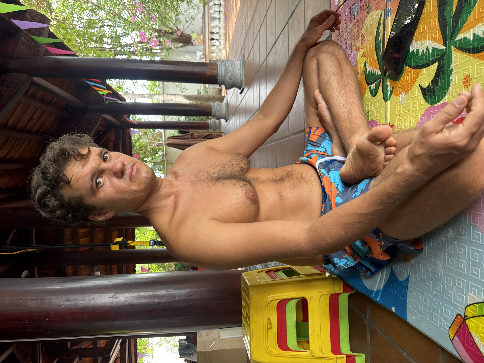 A man is depicted sitting cross-legged on a colorful, patterned mat in an outdoor or semi-outdoor setting. He is shirtless, revealing a tanned torso with some chest hair, and wears blue, orange, and black patterned swim trunks. His curly brown hair is styled casually, and he looks directly at the camera with a neutral to slightly serious expression.
The background features dark wooden pillars and beams, characteristic of traditional or rustic open-air architecture. Beyond these structures, green foliage and pink flowering trees are visible, suggesting a lush, tropical environment. On the mat beside the man's legs, a dark smartphone is placed, with its screen showing "28%." In the foreground, a stack of bright yellow, red, and green plastic stools can be seen.
The man's seated posture, with his hands resting on his knees, suggests a moment of relaxation or contemplation. The natural, bright lighting indicates it is daytime. The attire and the vibrant natural surroundings are consistent with warm weather and a leisure context, such as a resort or guesthouse in Mũi Né, Vietnam.